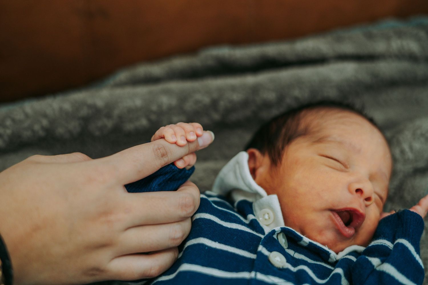 Newborn baby yawning while grasping parent's finger, wearing blue striped outfit on gray blanket.