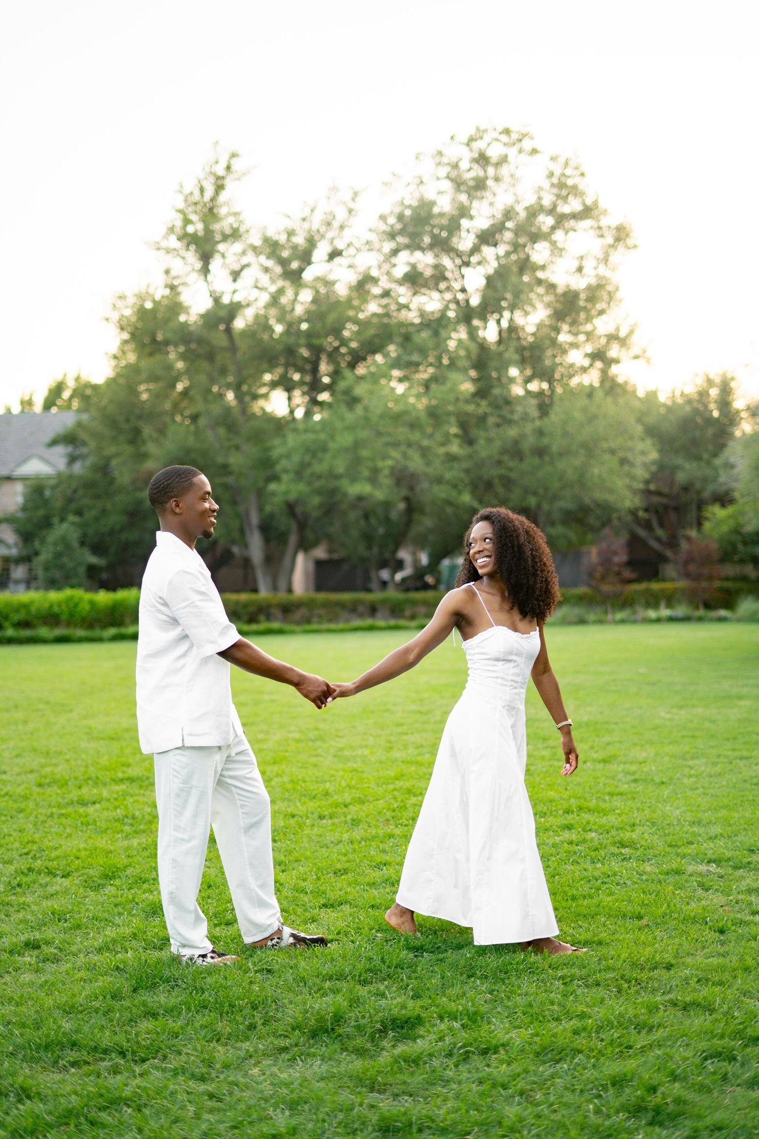 Joyful Black couple in white outfits holding hands and laughing together on a lush green lawn.