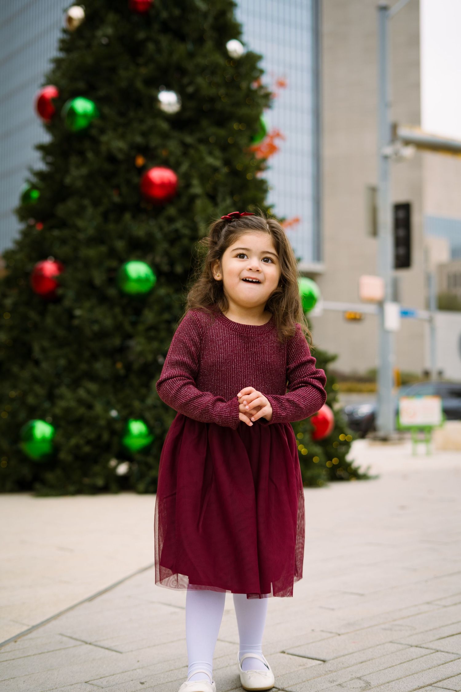 Adorable little girl in burgundy dress smiles joyfully while standing in front of a large Christmas tree.