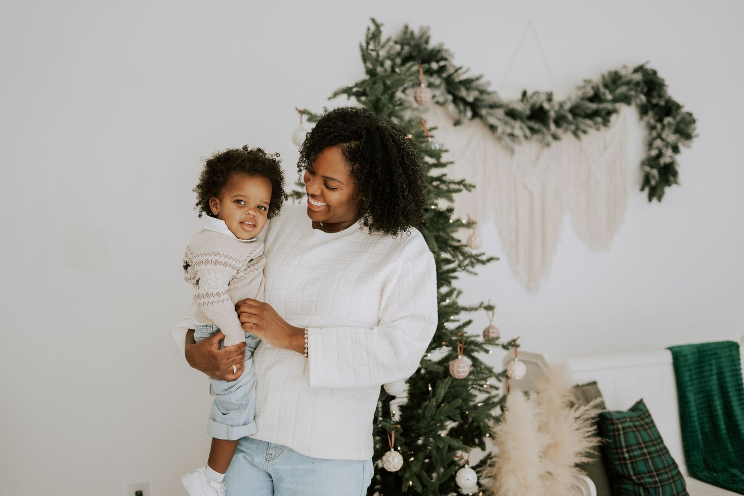 Smiling mother in white sweater holds toddler near decorated Christmas tree indoors.