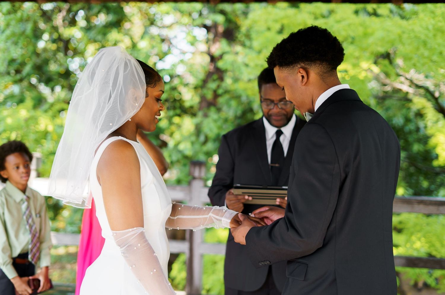 Groom places wedding ring on bride's finger during intimate outdoor ceremony under a lush green pavilion.
