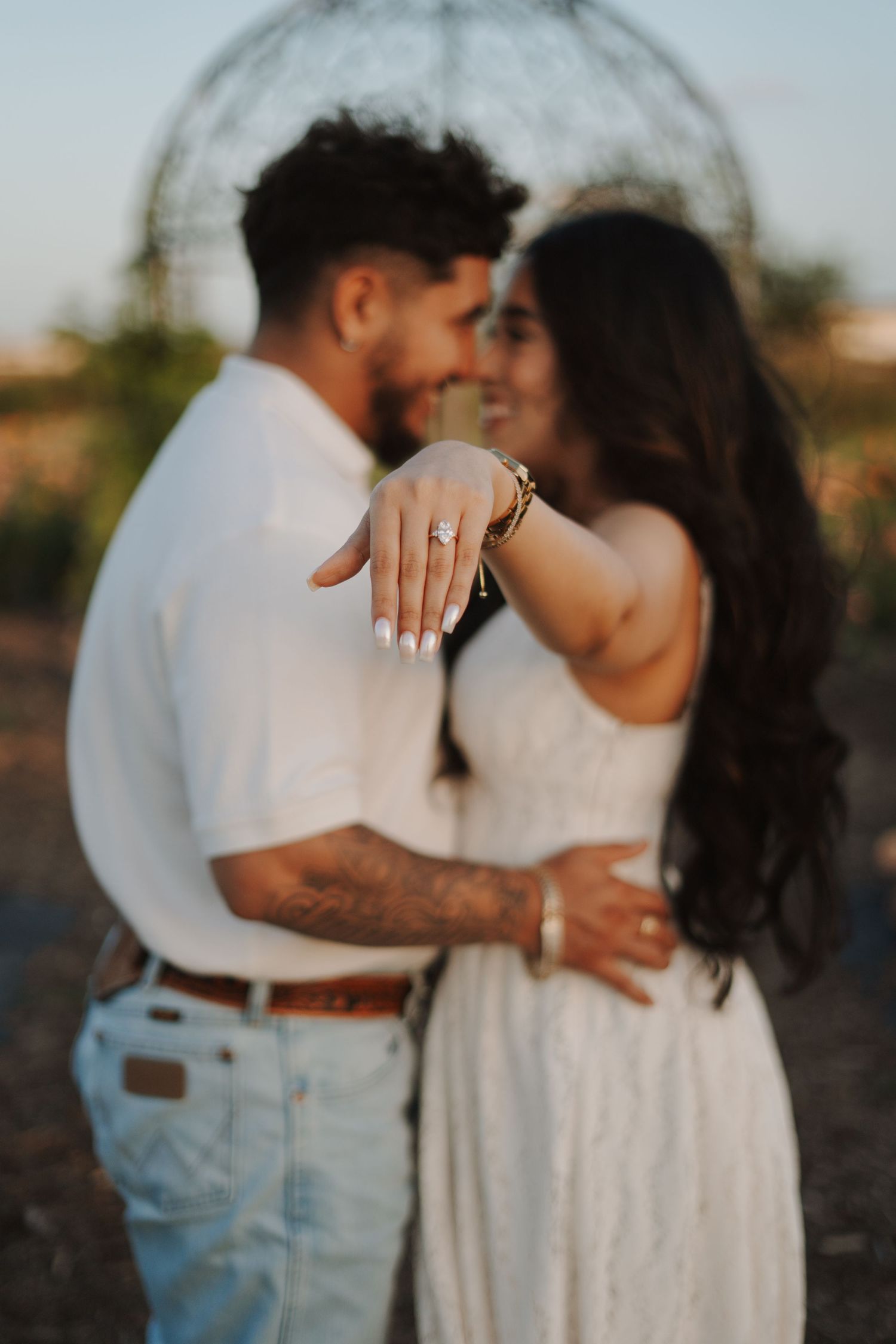 Couple in white embraces outdoors, woman showing diamond engagement ring, bokeh background with garden arch.