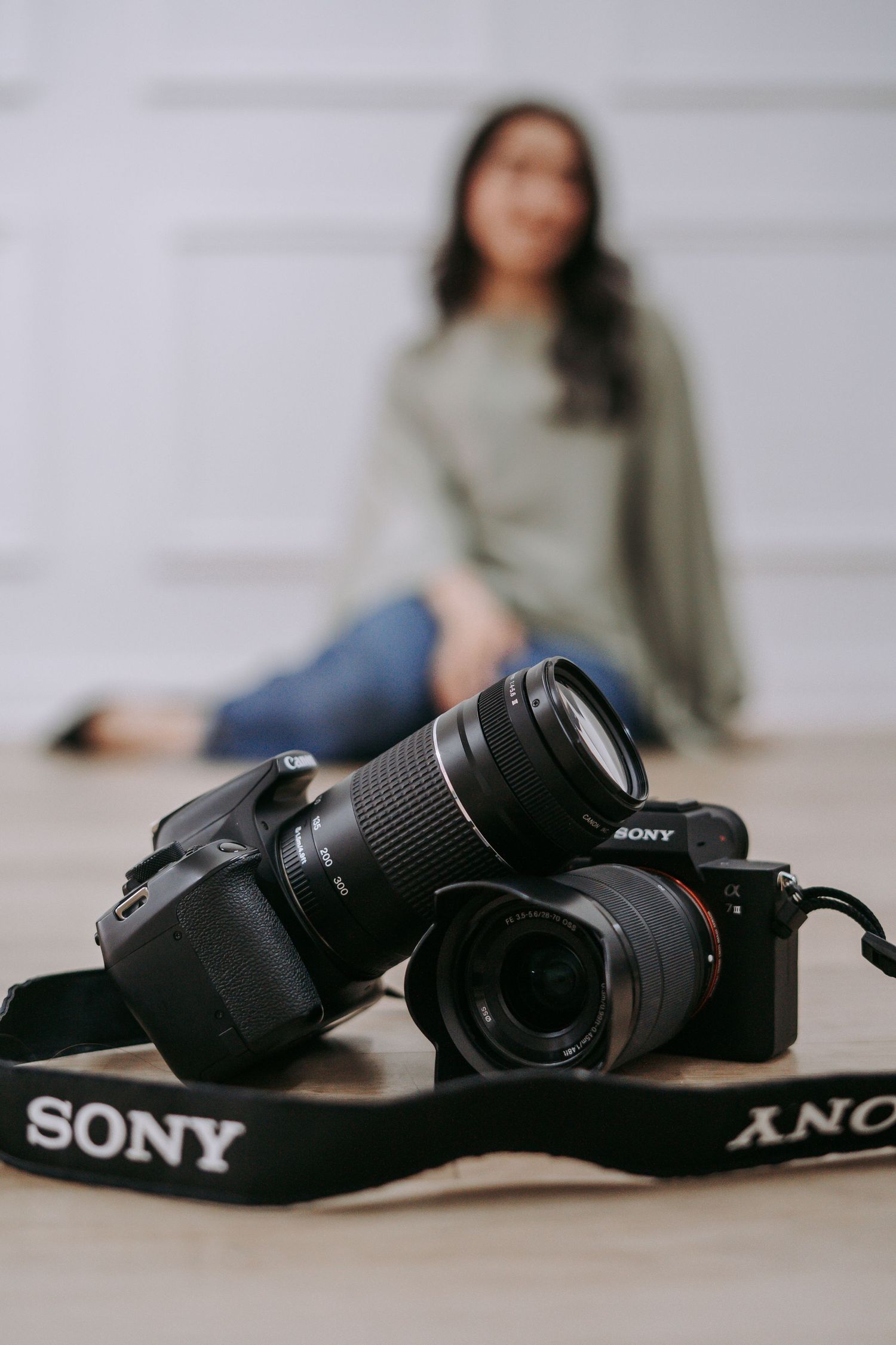 Two Sony DSLR cameras with lenses on wooden floor with blurred woman sitting in background.