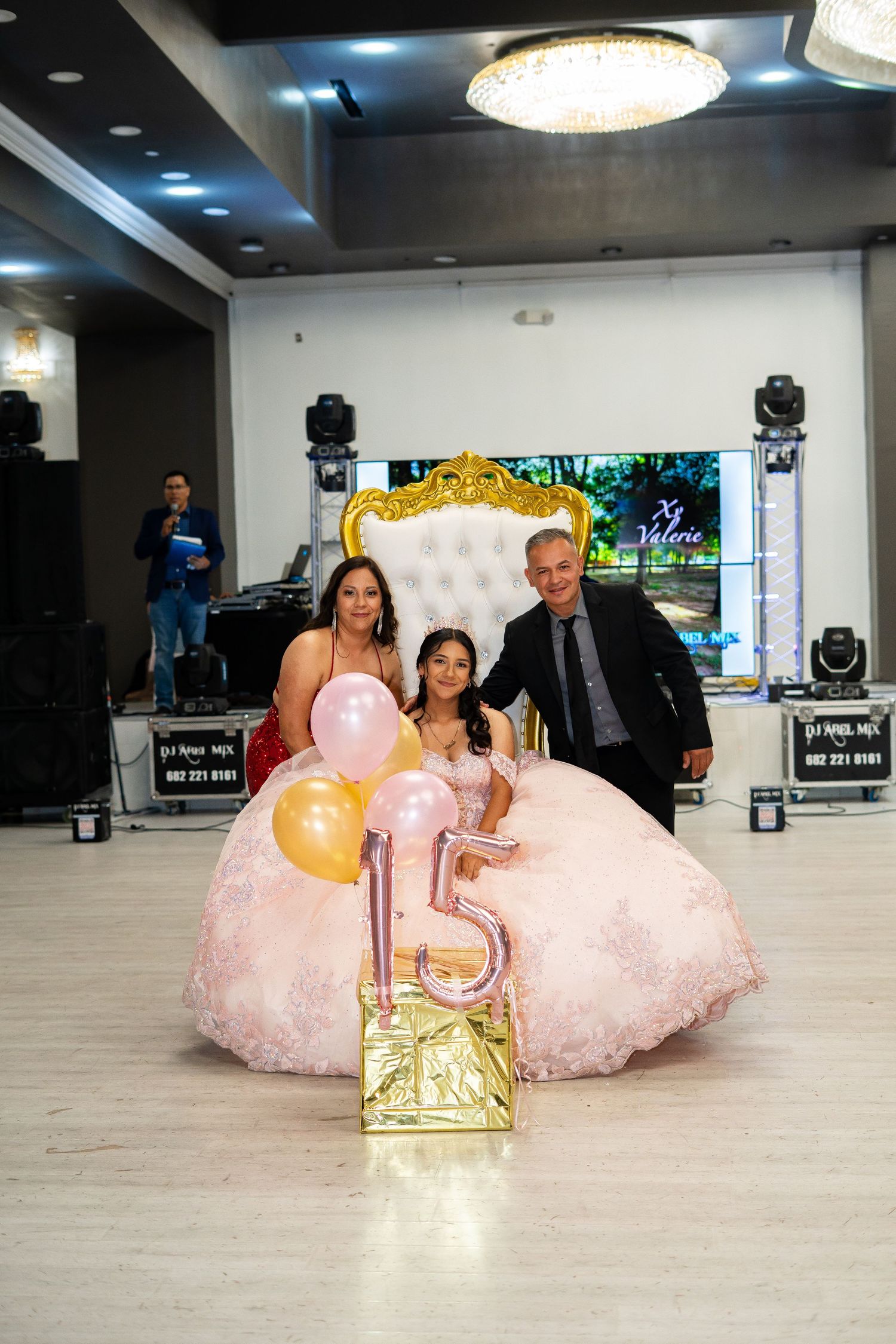 A quinceañera poses with her parents beside number 15 balloons at her party.