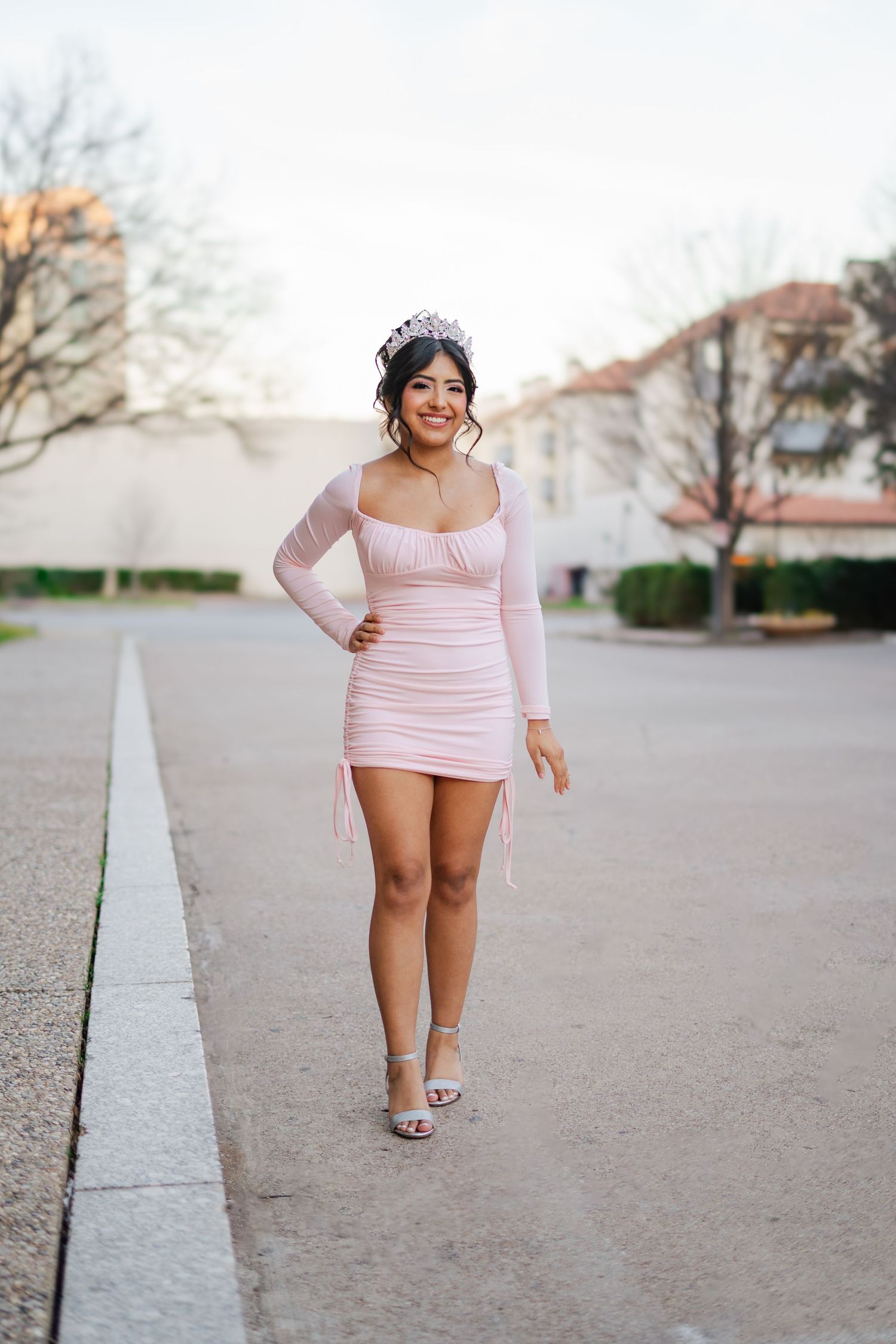 Young Latina woman in pink mini dress and tiara crown poses outdoors at sunset for quinceañera photos.