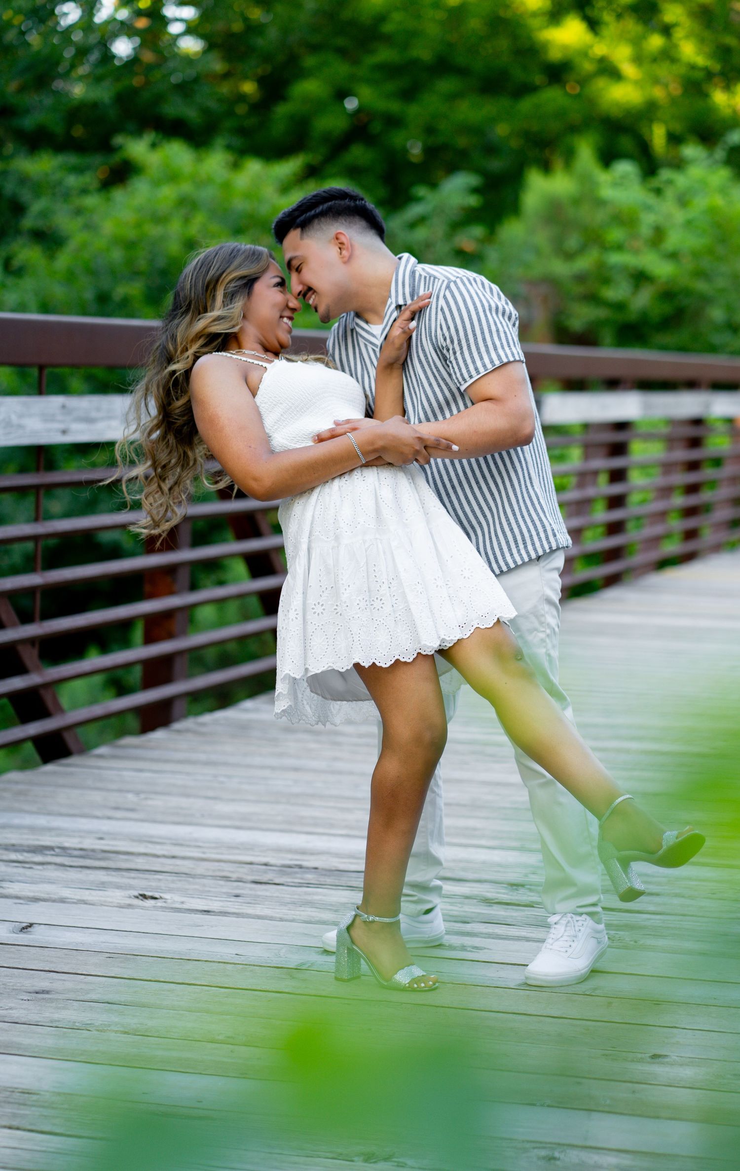 Couple shares romantic moment on wooden bridge surrounded by lush green foliage.