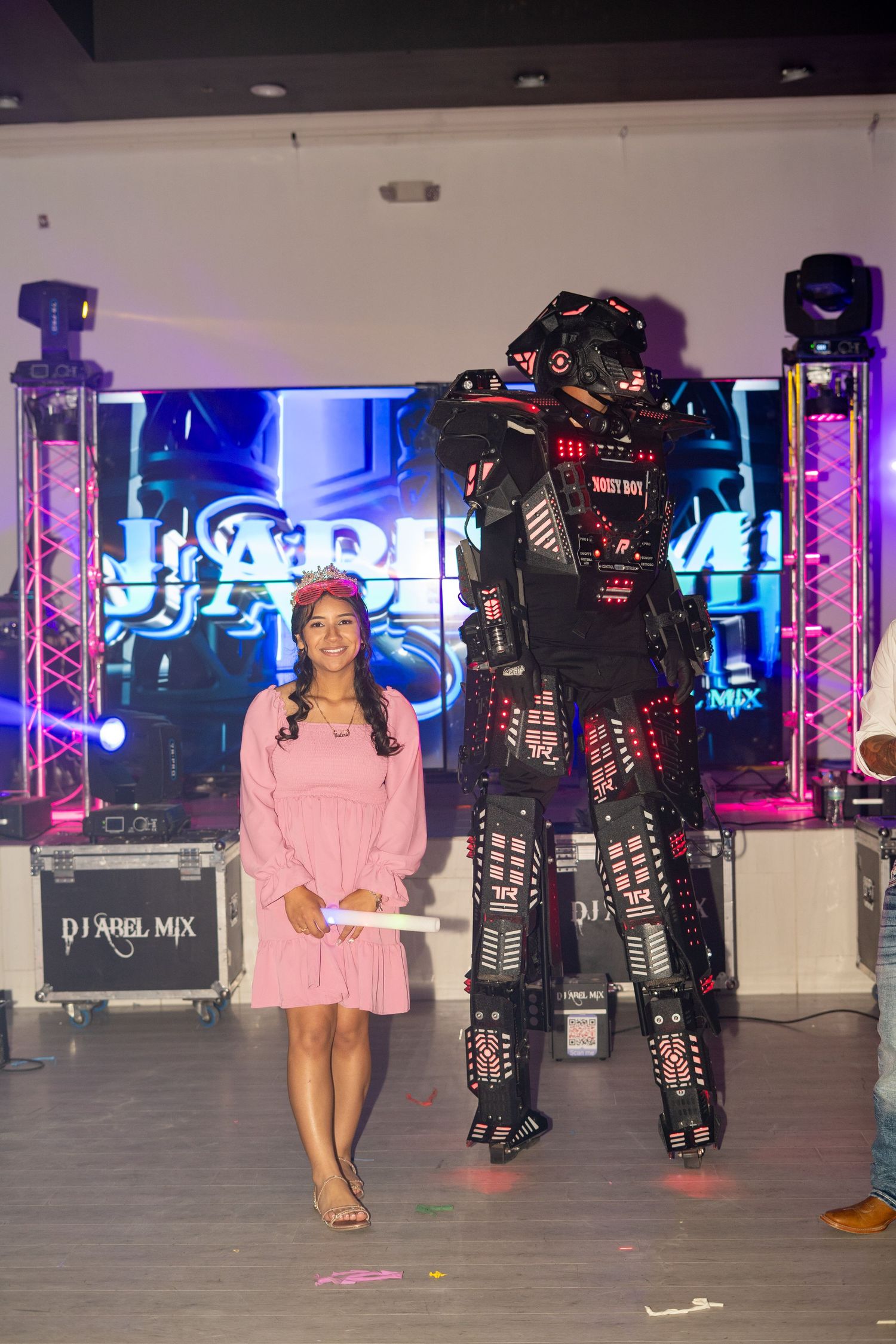 A young woman in pink dress poses beside an illuminated robot costume at a quinceañera party.