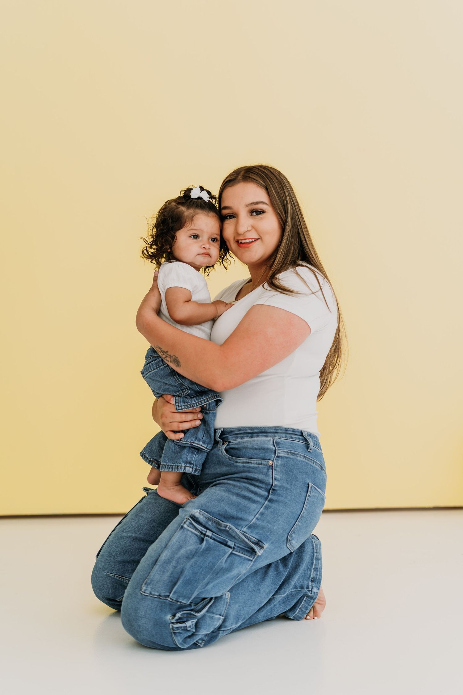 Young woman in white tee and jeans kneeling on floor, lovingly holding a baby girl in denim outfit.