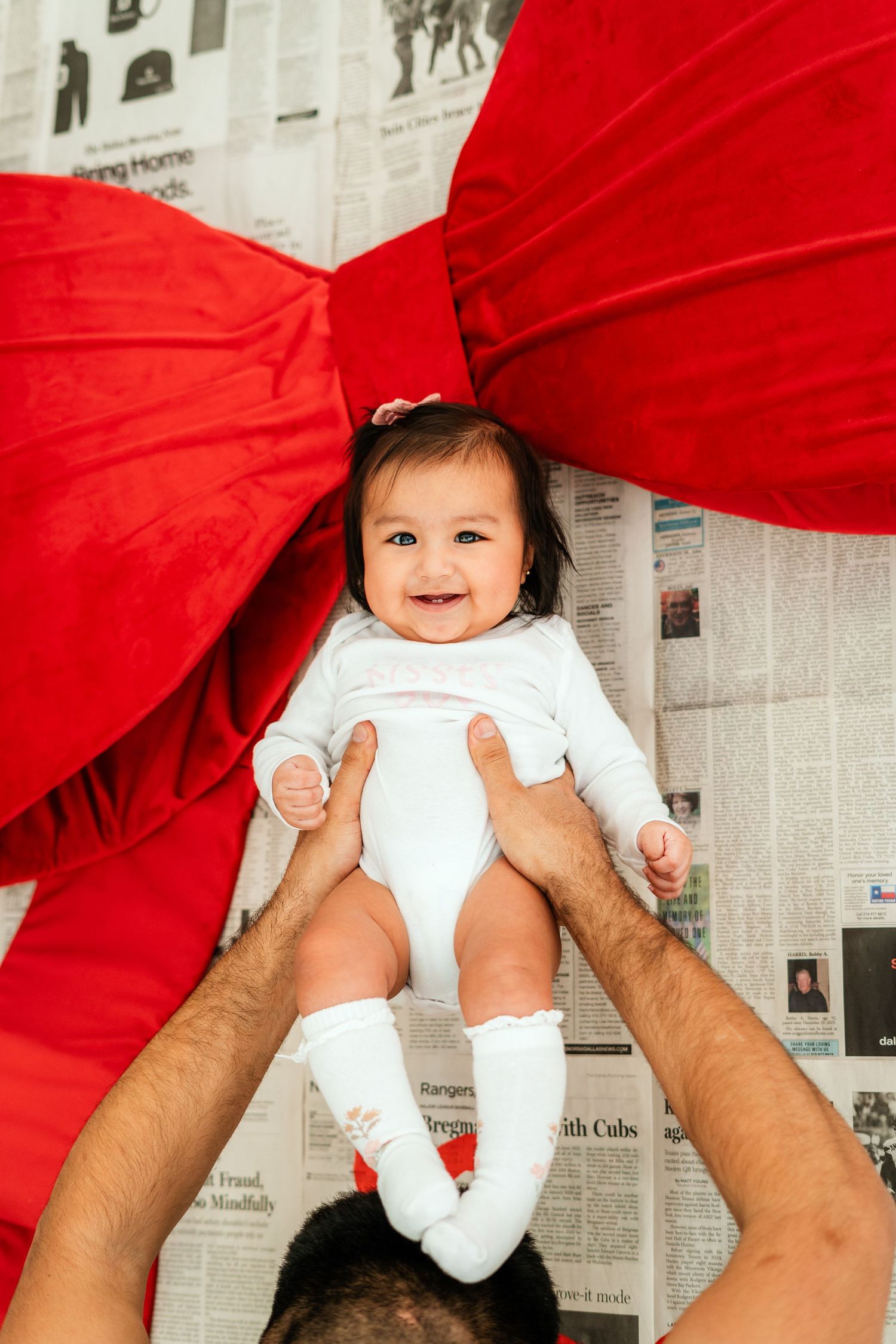 Joyful baby in white onesie held up by father beneath large red bow on newspaper backdrop.