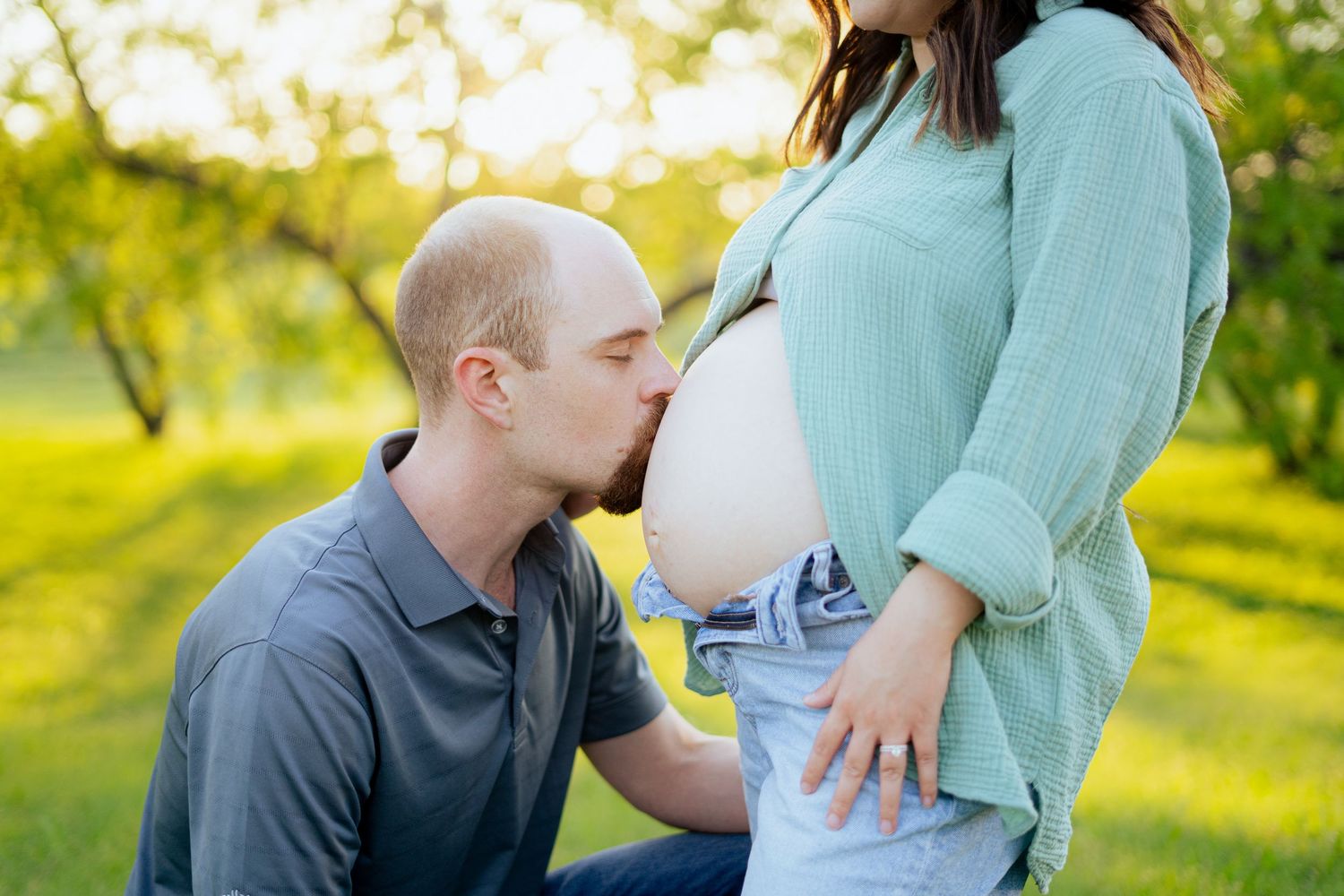 Man kneeling to kiss pregnant woman's bare baby bump outdoors in warm golden sunlight during maternity session.