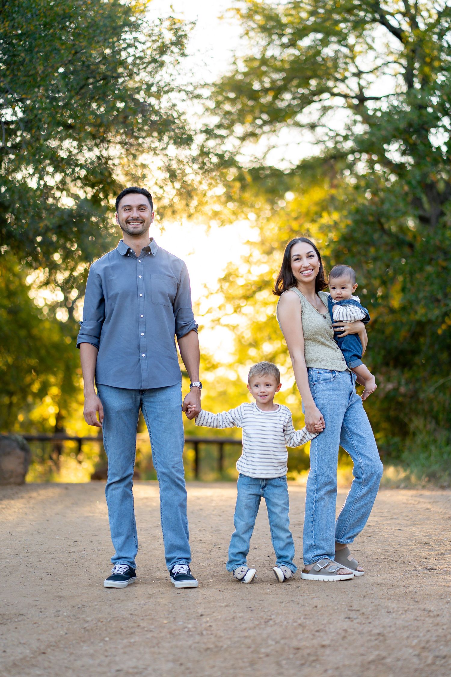 Family of four holding hands on a sunlit dirt path surrounded by golden autumn trees.