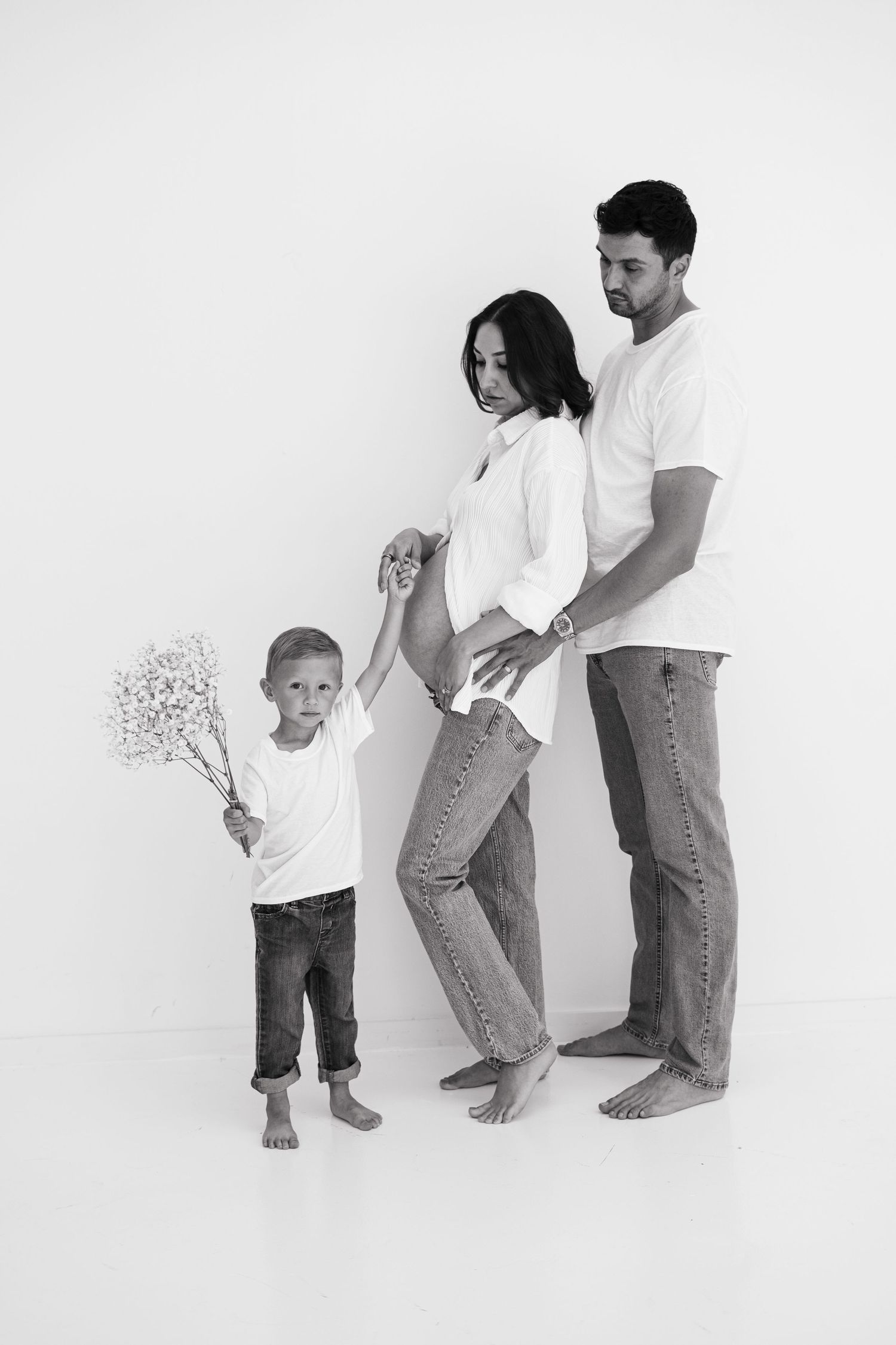Black and white maternity photo of a family in white shirts and jeans with a young boy holding flowers.