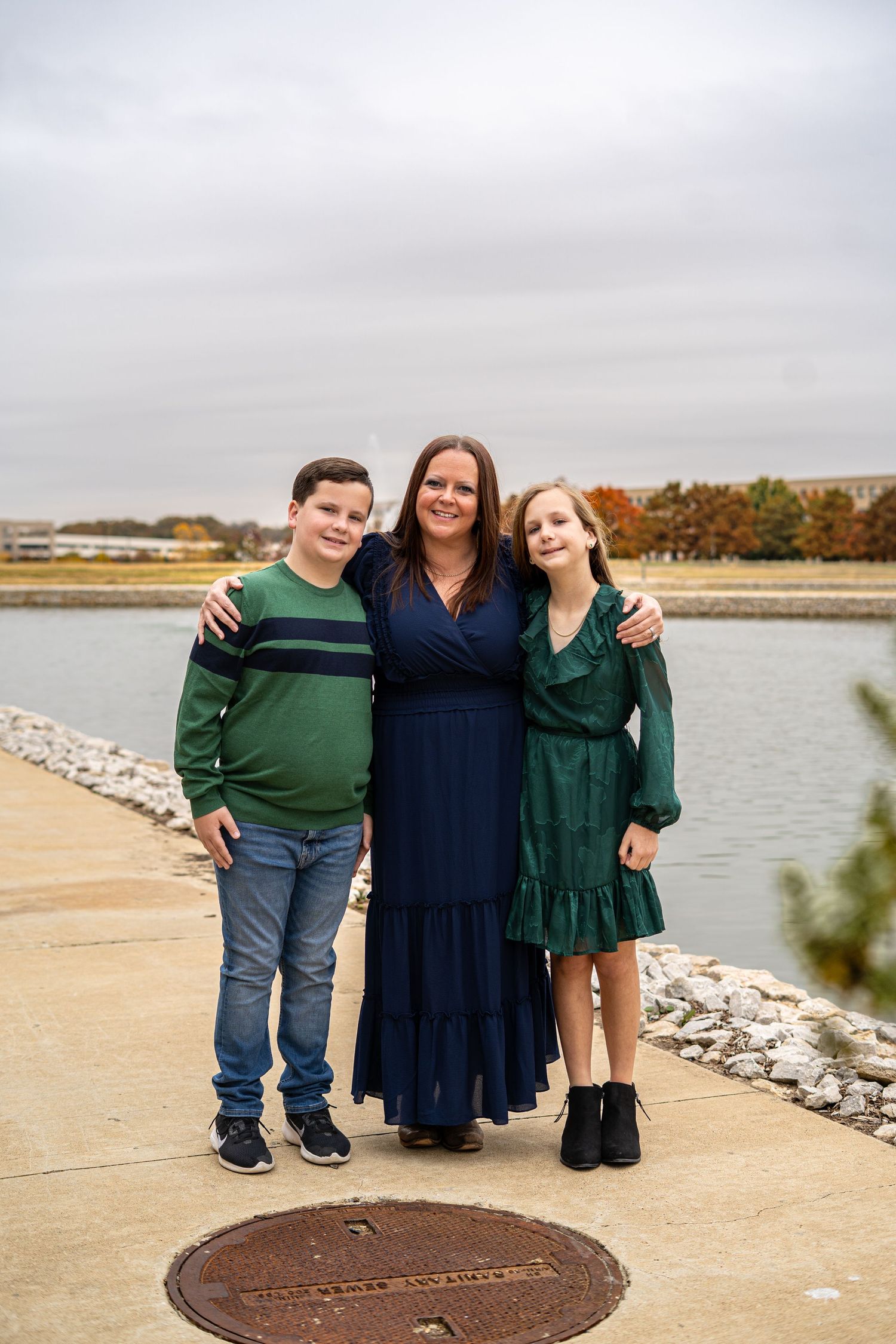 A woman in a navy dress poses with two children by a peaceful waterfront on an overcast day.