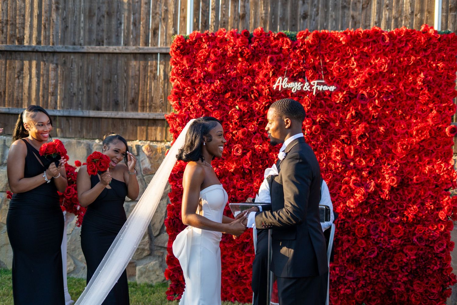Bride and groom stand at altar with bridesmaids in black dresses beside vibrant red rose flower wall.