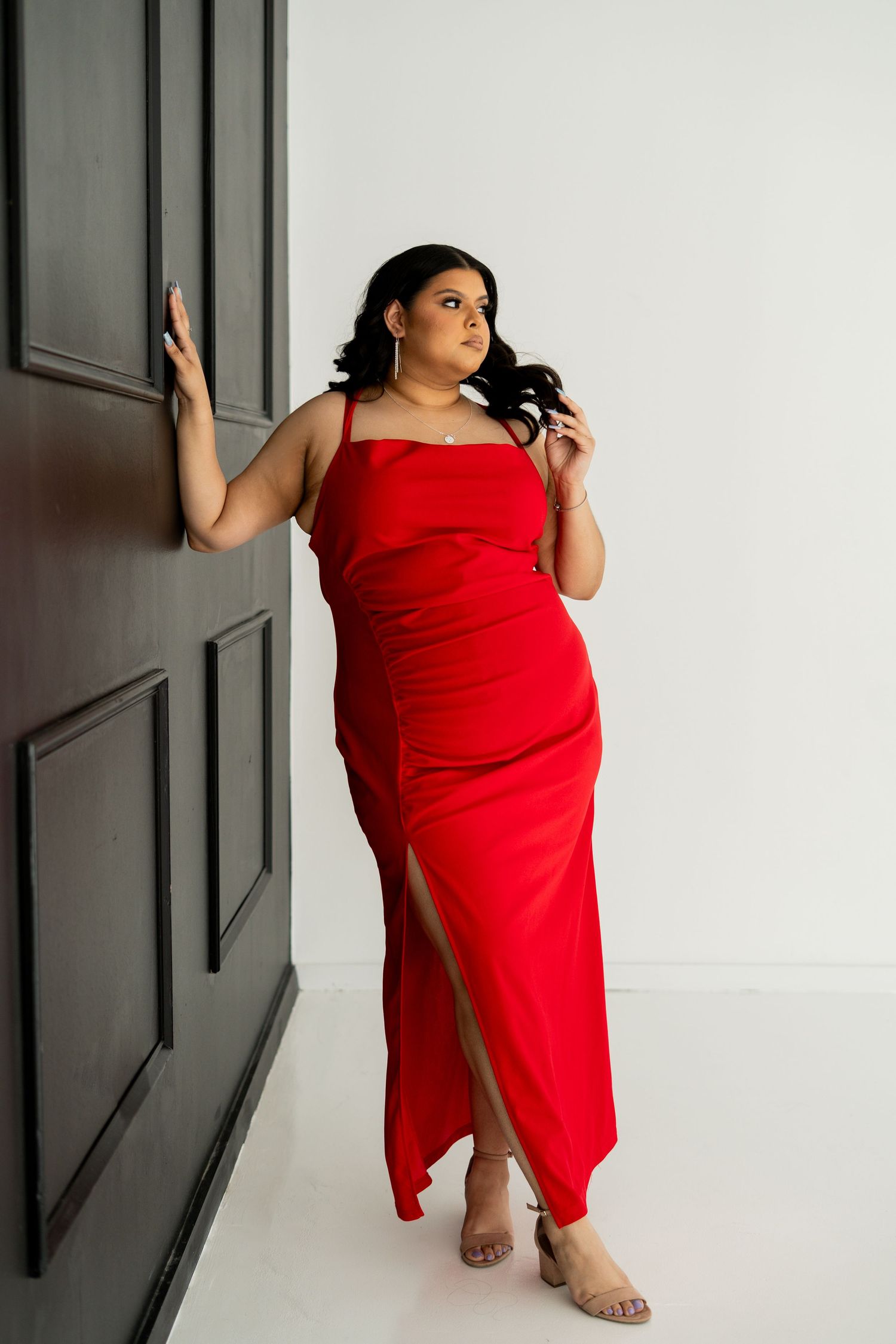 Woman in red sleeveless gown leaning against dark door looking away in studio.