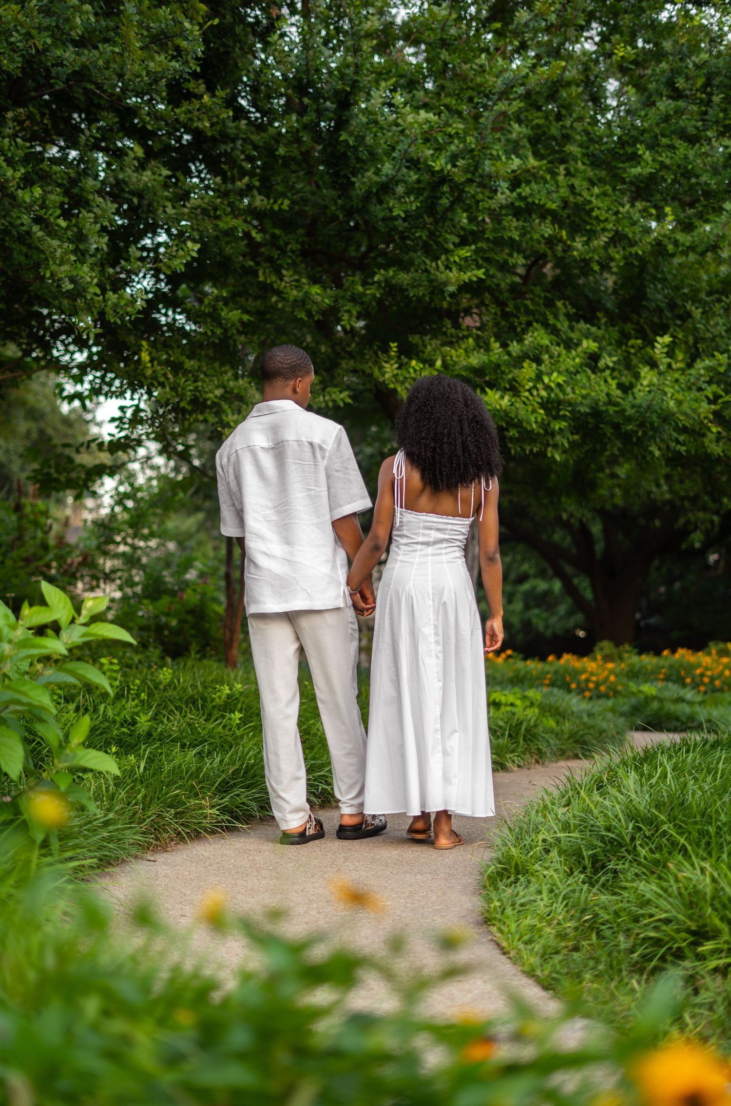 A Black couple in white outfits walks hand-in-hand away from camera along a lush green garden path.