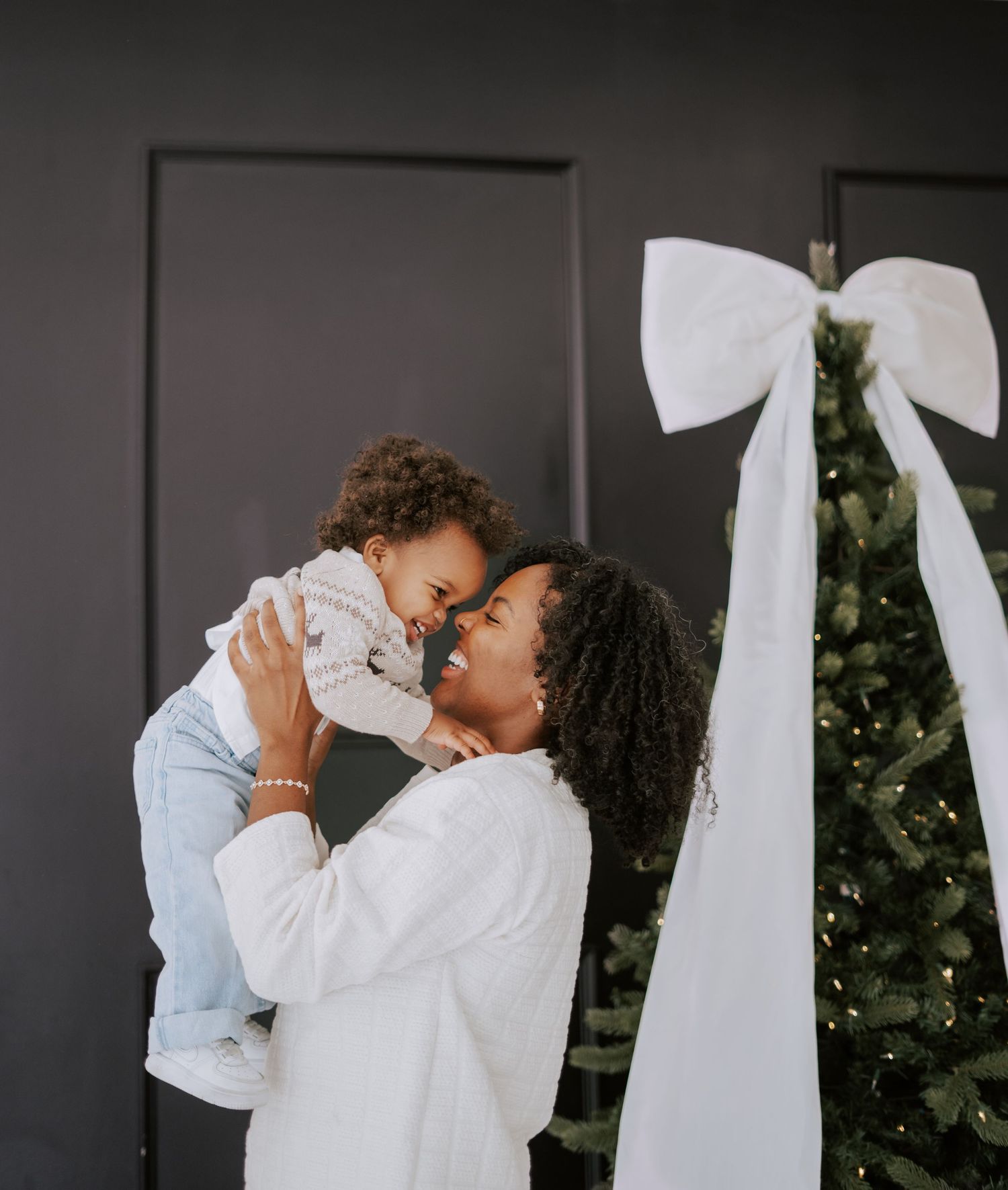 A loving mother lifts her baby up for a kiss beside a Christmas tree decorated with a white bow.
