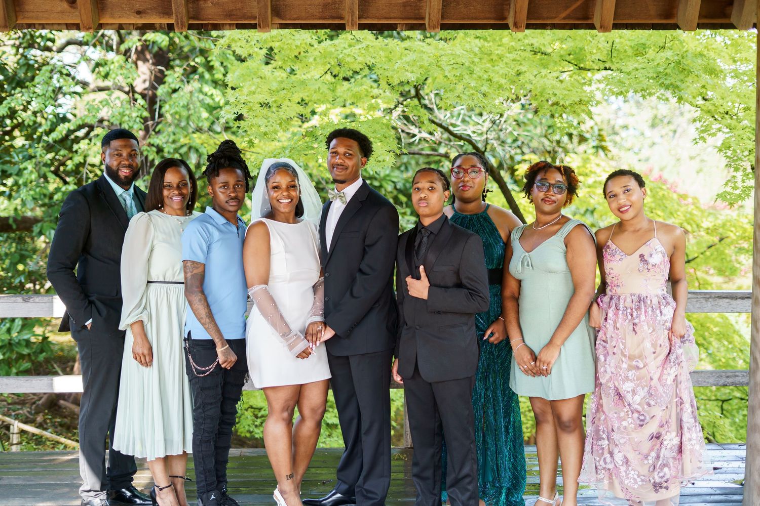 Large wedding party group photo with nine guests posing under a gazebo surrounded by lush greenery.