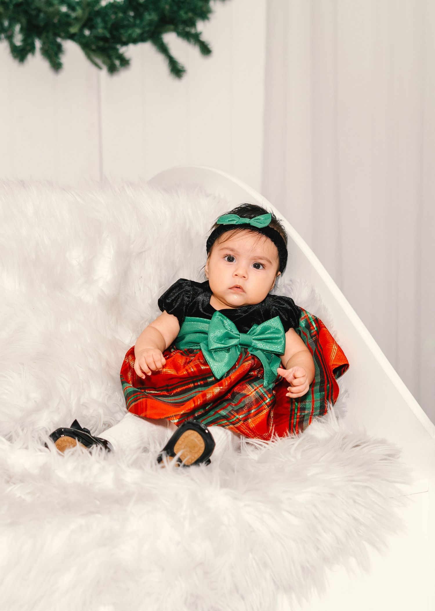 Adorable baby in festive red and green plaid dress with green bow headband sits on white fur blanket.