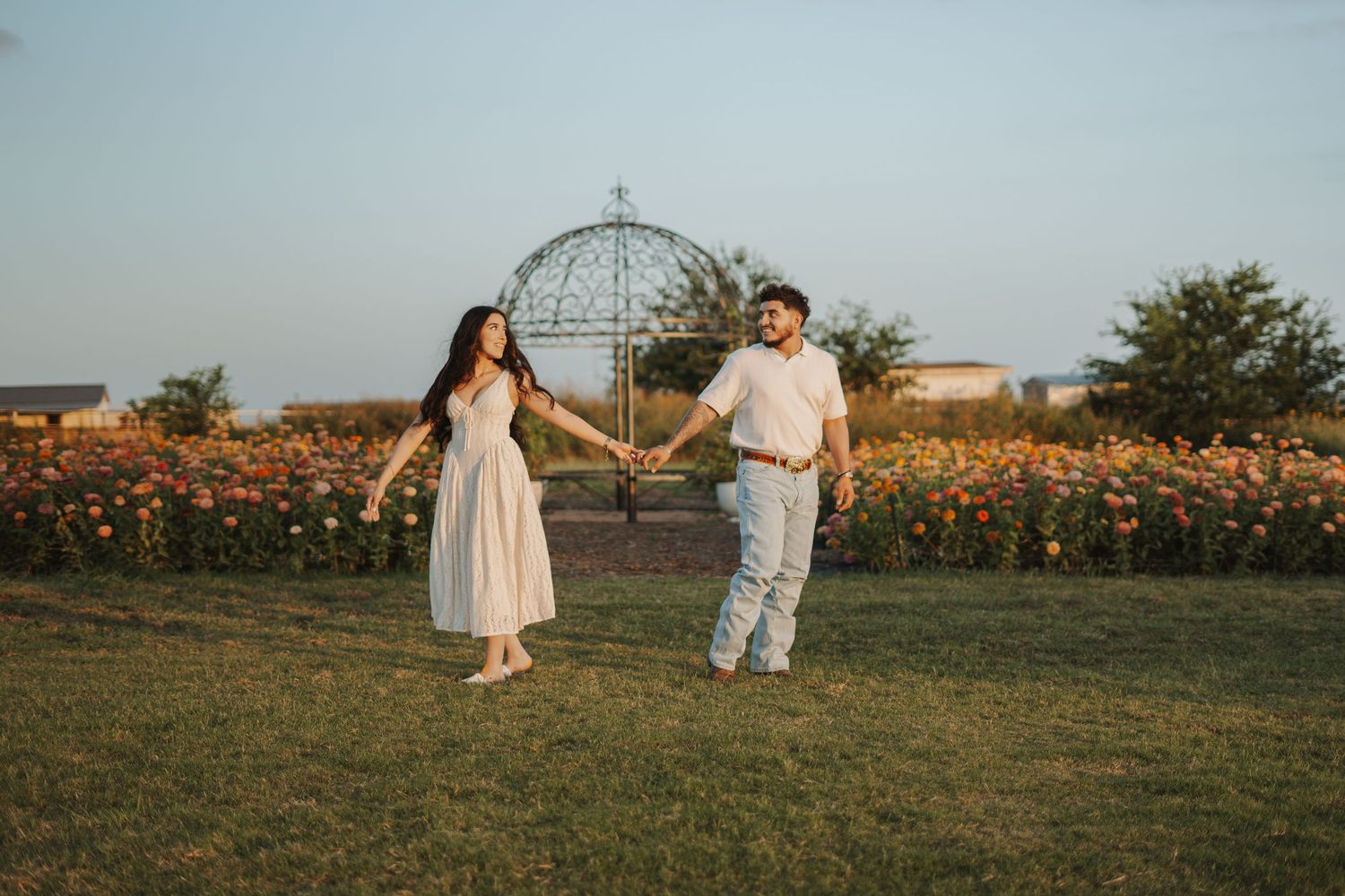 Couple holding hands walking in a garden with colorful flowers and a decorative metal globe sculpture at golden hour.