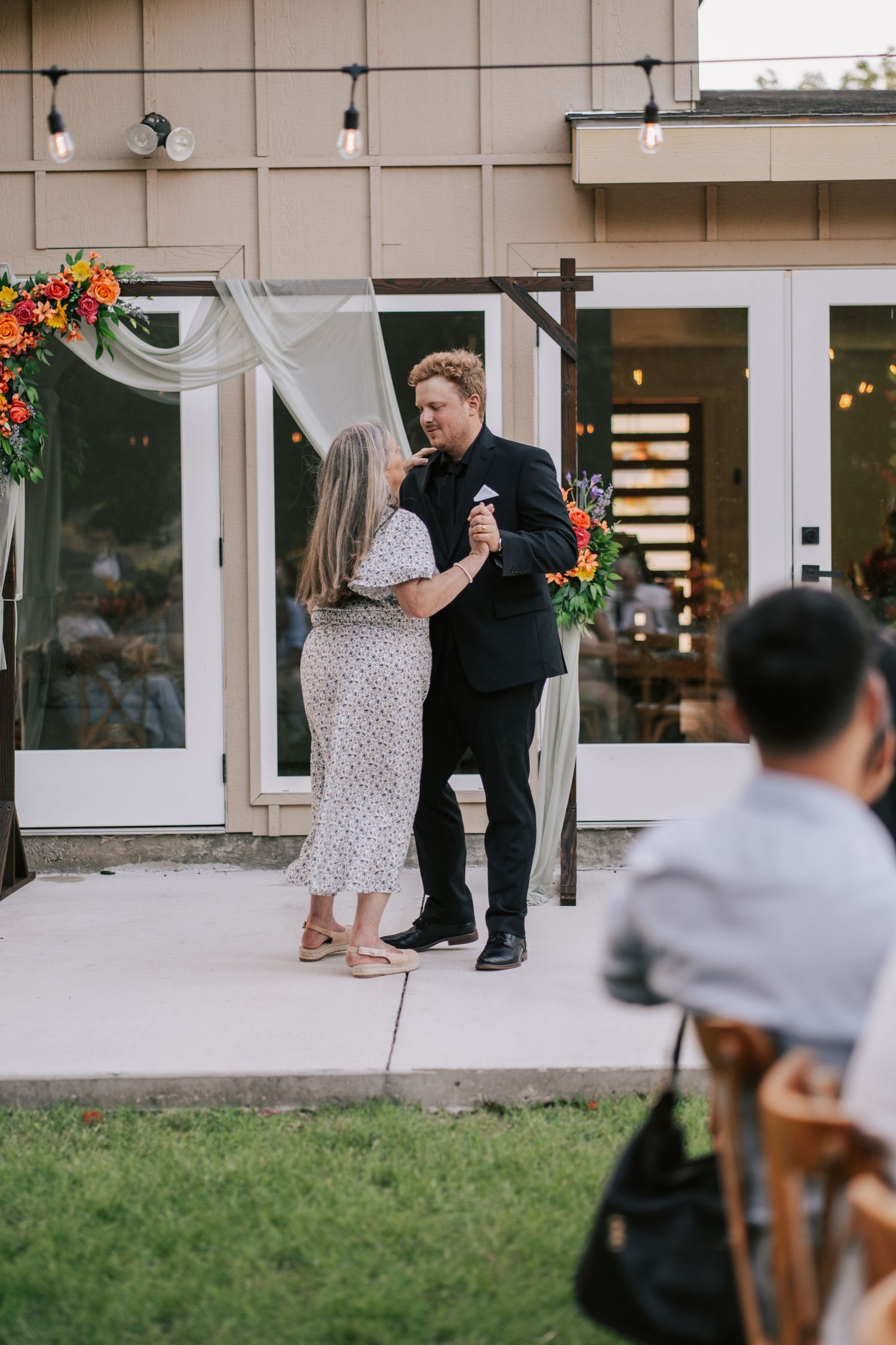 Groom in black suit shares a dance with a woman in a floral dress at an outdoor wedding reception.