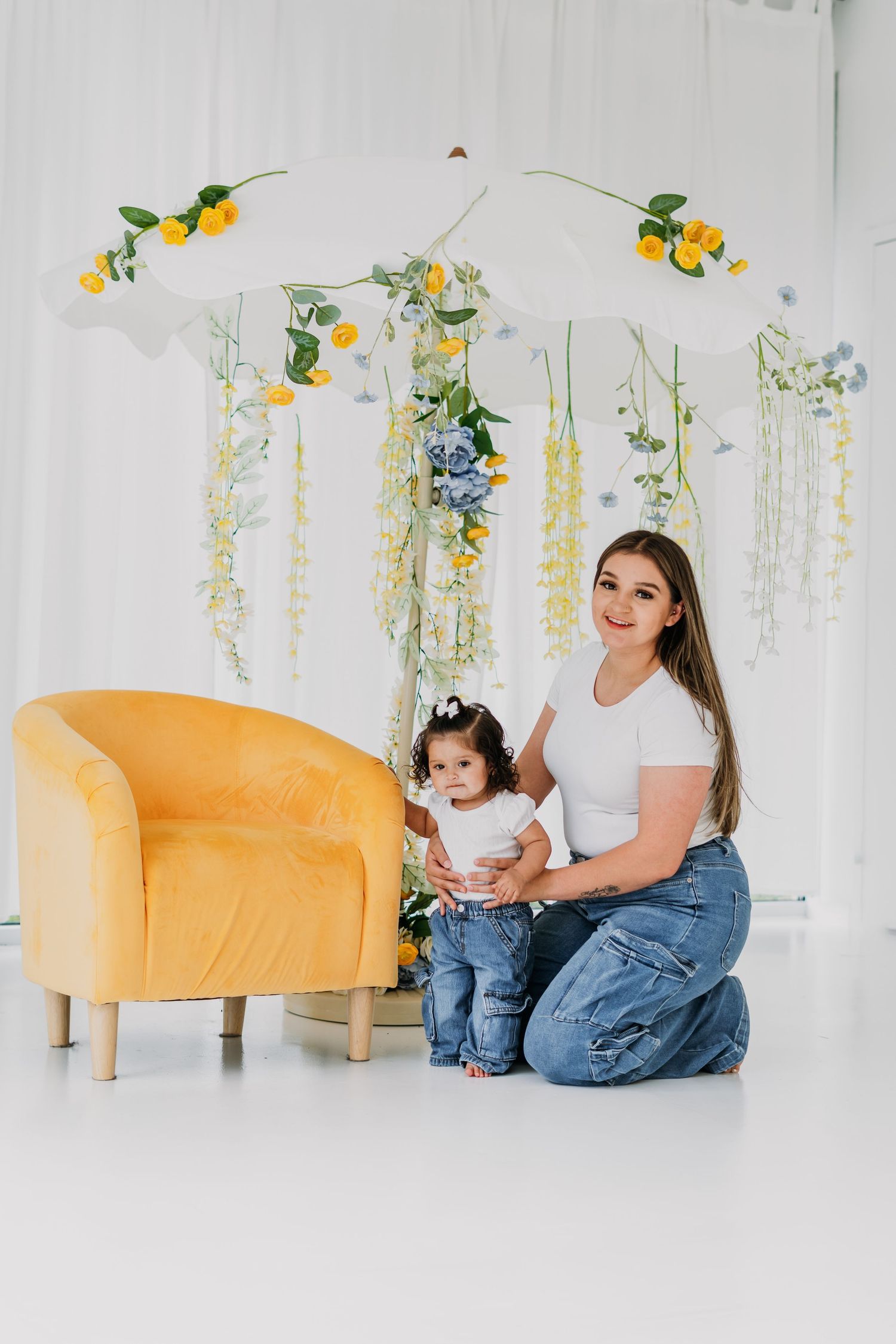 A mother and baby girl pose together on the floor beside a yellow velvet chair with floral spring decor backdrop.