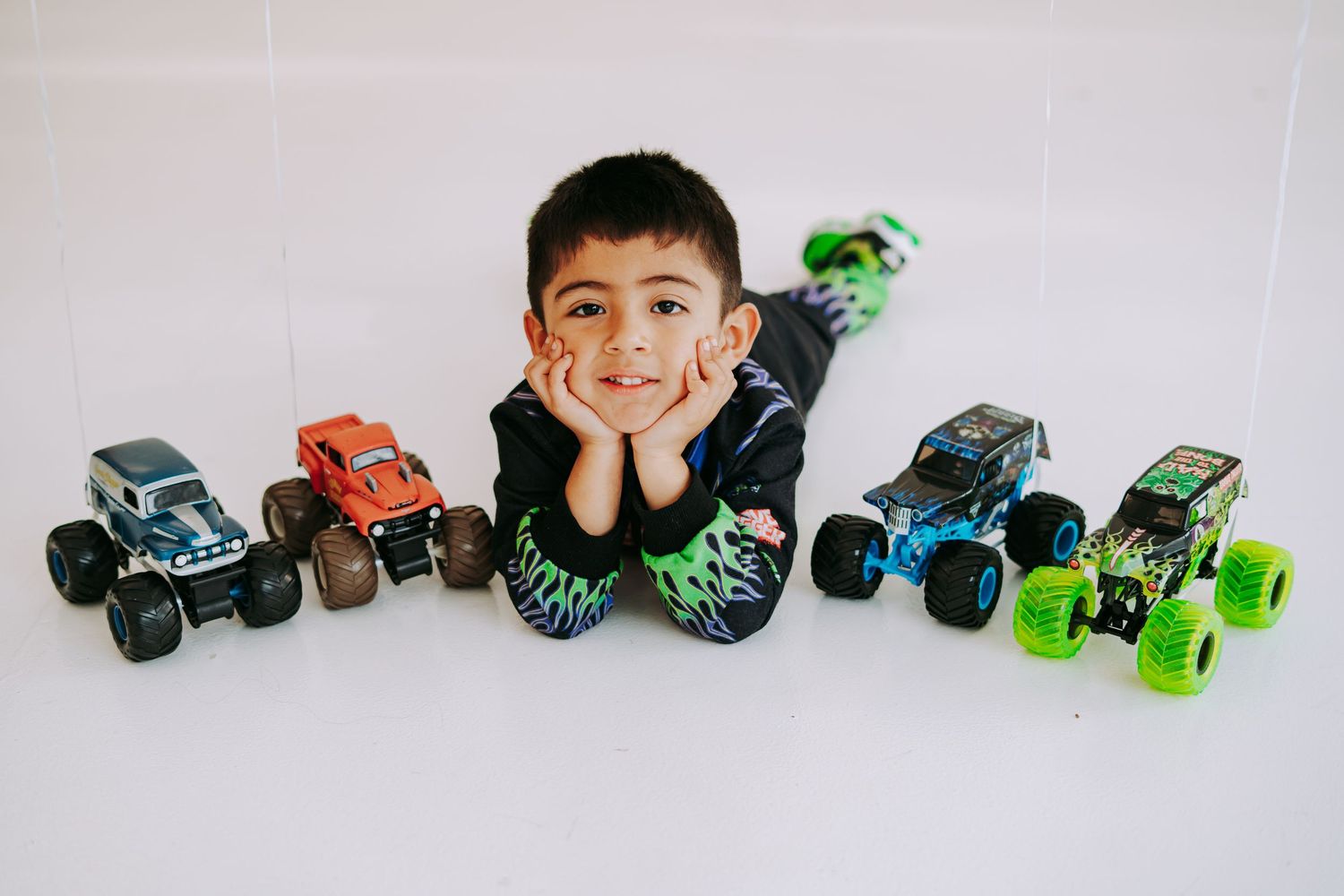 Young boy lying on floor surrounded by colorful monster trucks toy collection.