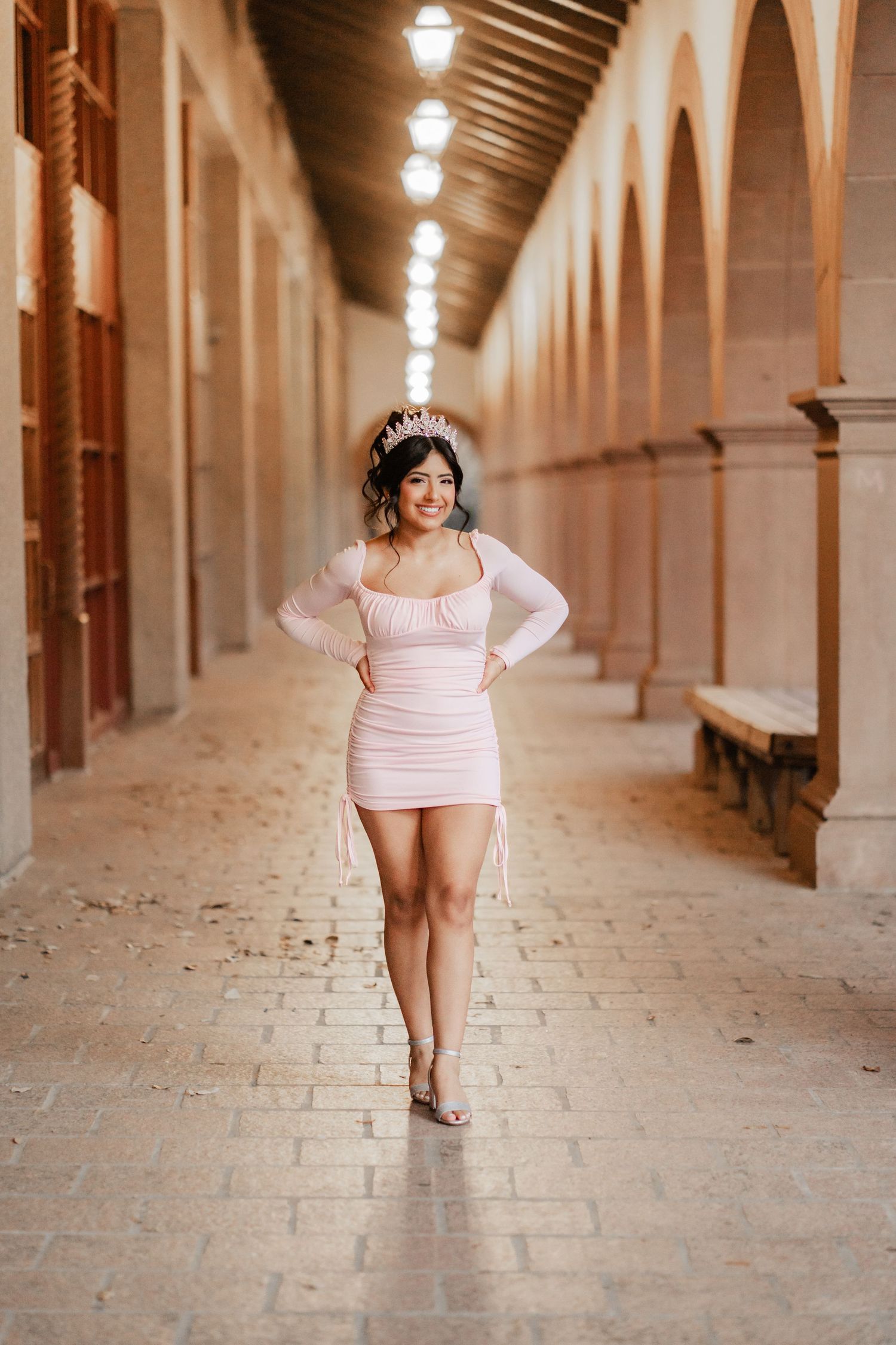 Young Latina in pink mini dress and tiara walks confidently through an elegant arched corridor.