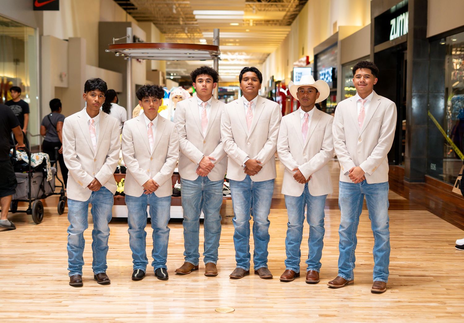 Six young men in white blazers and jeans posing together inside a shopping mall corridor.