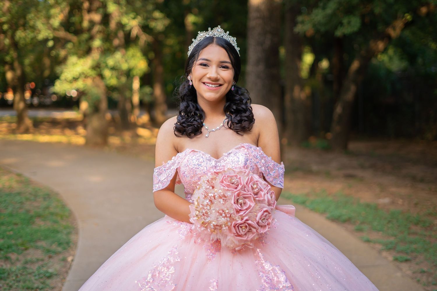Smiling young woman in a pink quinceañera gown holding a rose bouquet, wearing a tiara in a park.