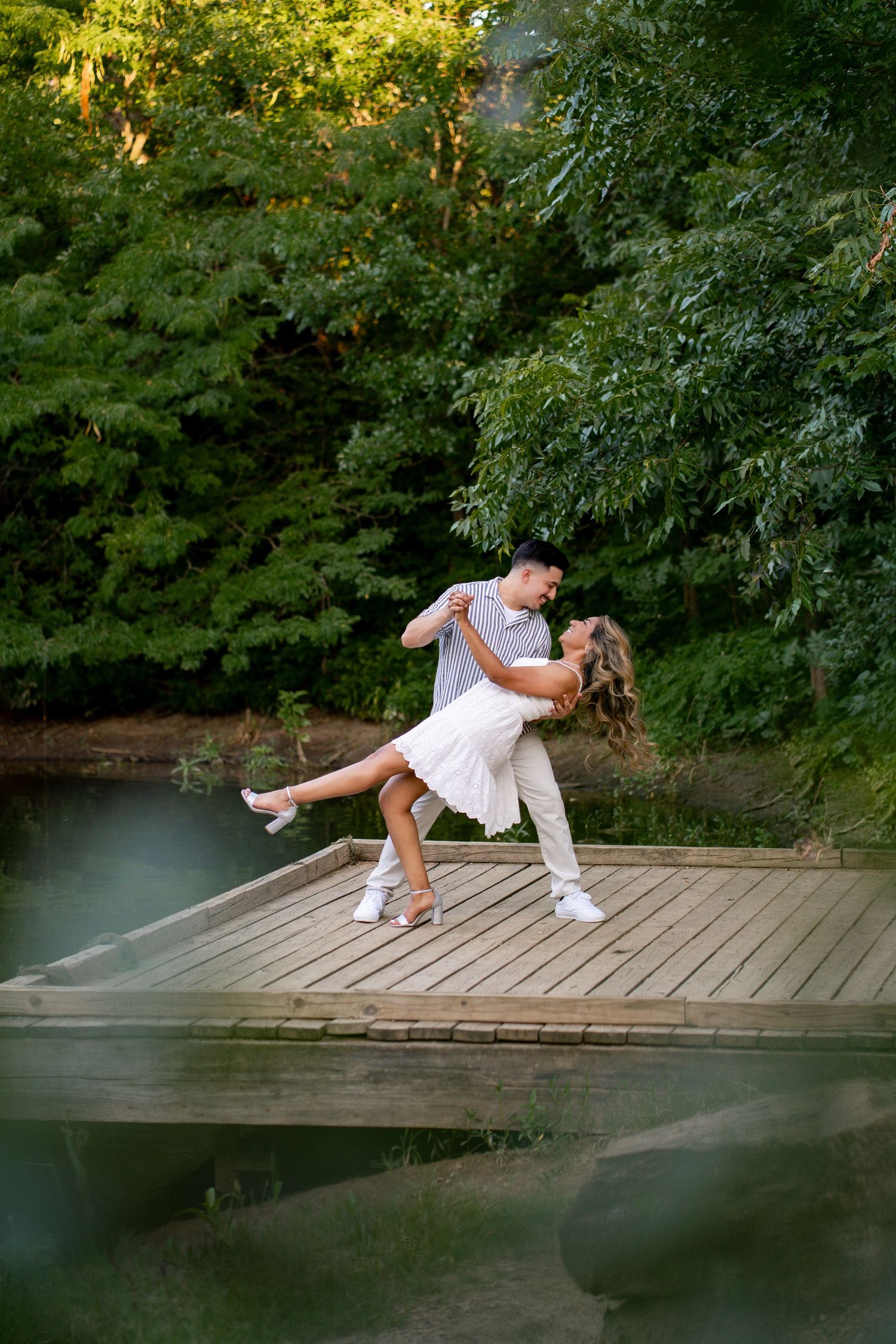 Man dipping woman in a white dress on a wooden dock surrounded by lush green trees.