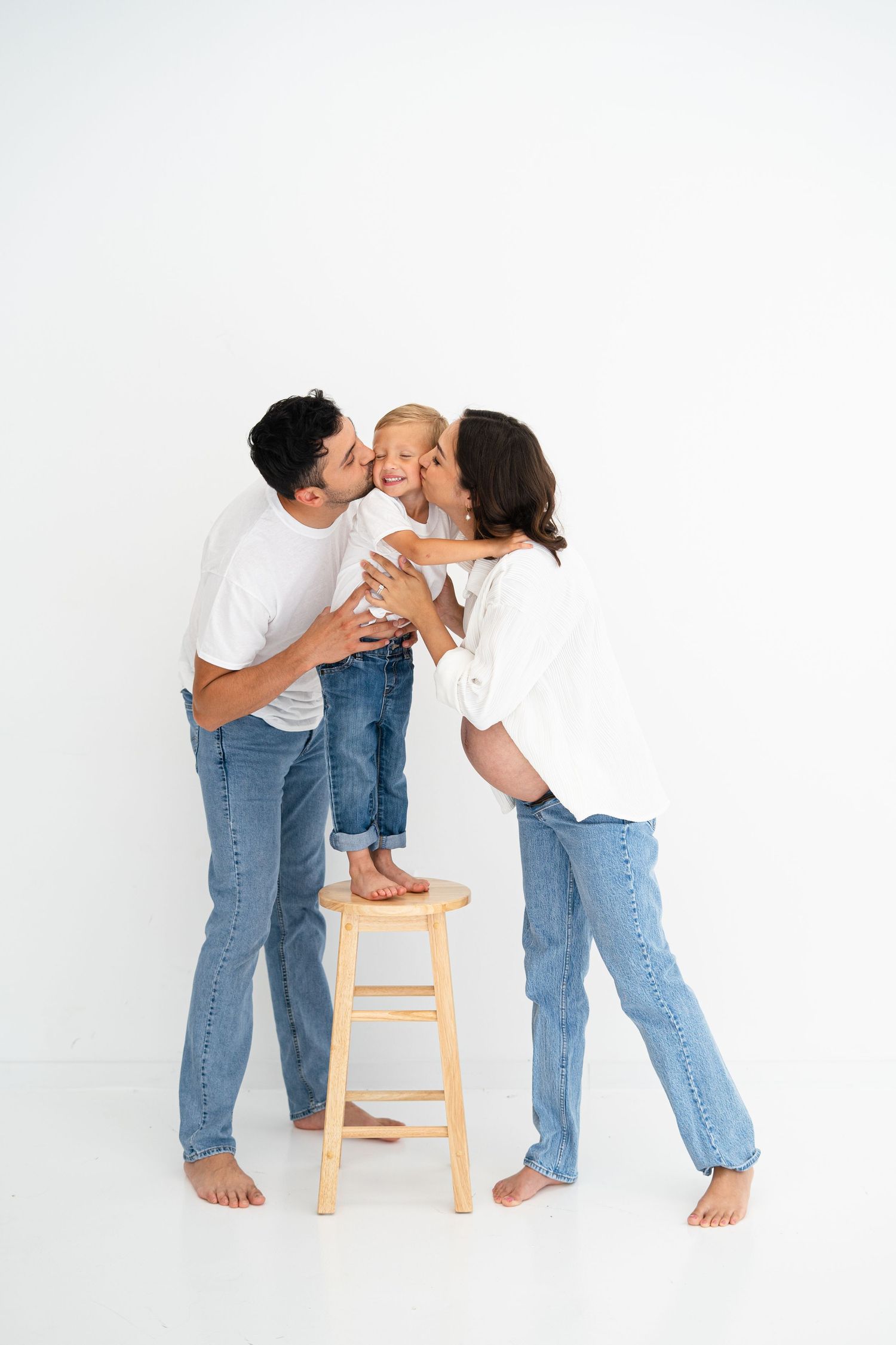 Parents kissing their young child standing on wooden stool in white and denim outfits.