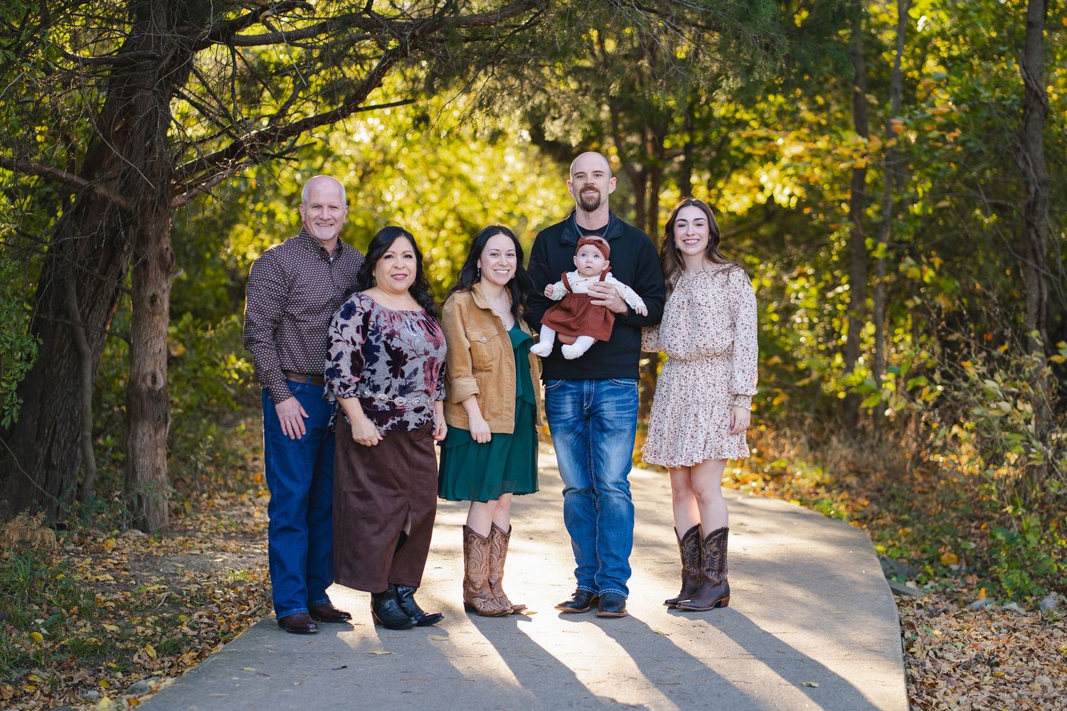 Family of six posing on a tree-lined path during autumn, wearing coordinated fall outfits with a baby in the center.