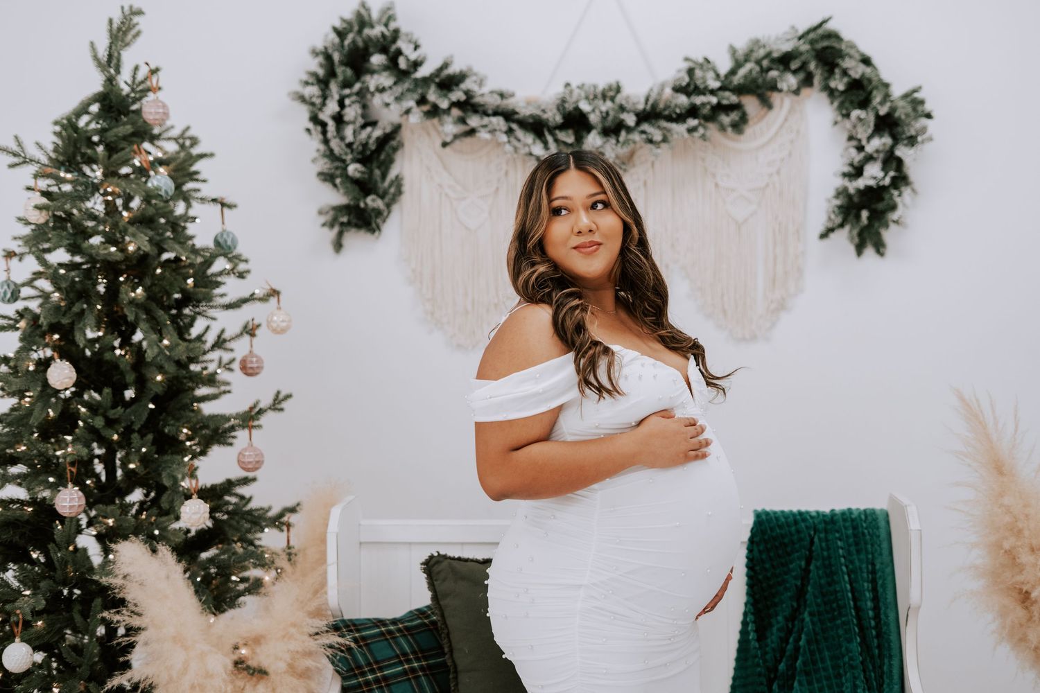 Pregnant woman in white off-shoulder gown poses beside Christmas tree with macrame wall hanging decor.
