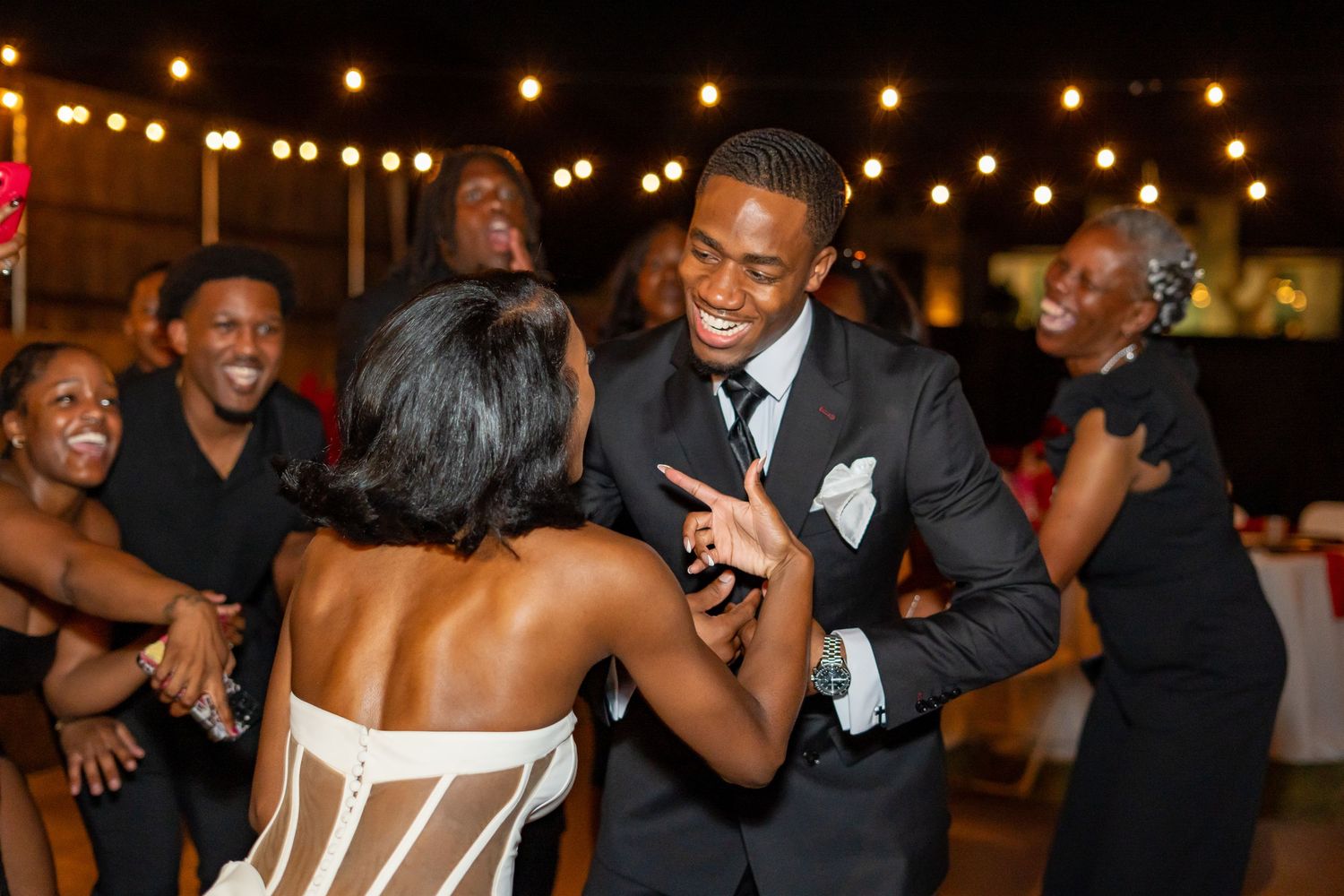 Groom in black suit smiles while dancing with bride at wedding reception under string lights.