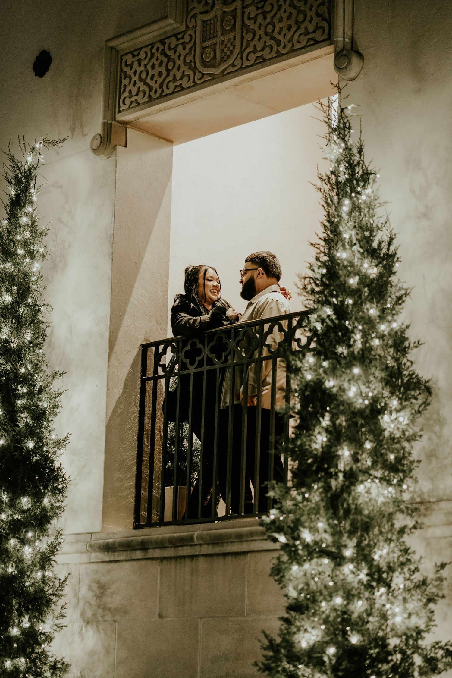 Romantic couple sharing a tender moment on an ornate balcony surrounded by twinkling Christmas trees at night.