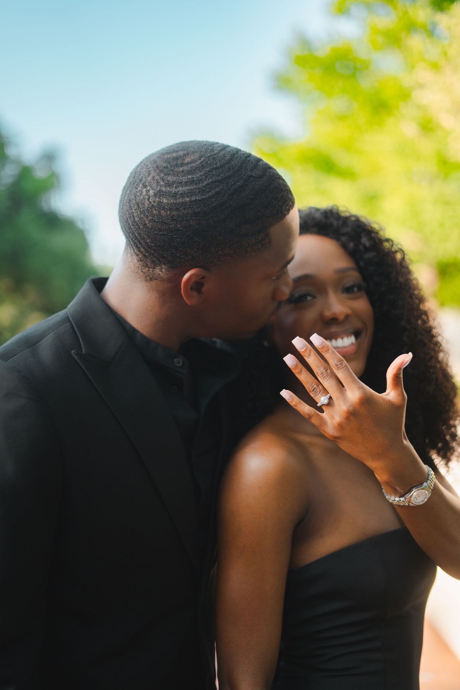 Newly engaged Black woman smiling and showing off her ring while her partner kisses her cheek outdoors.