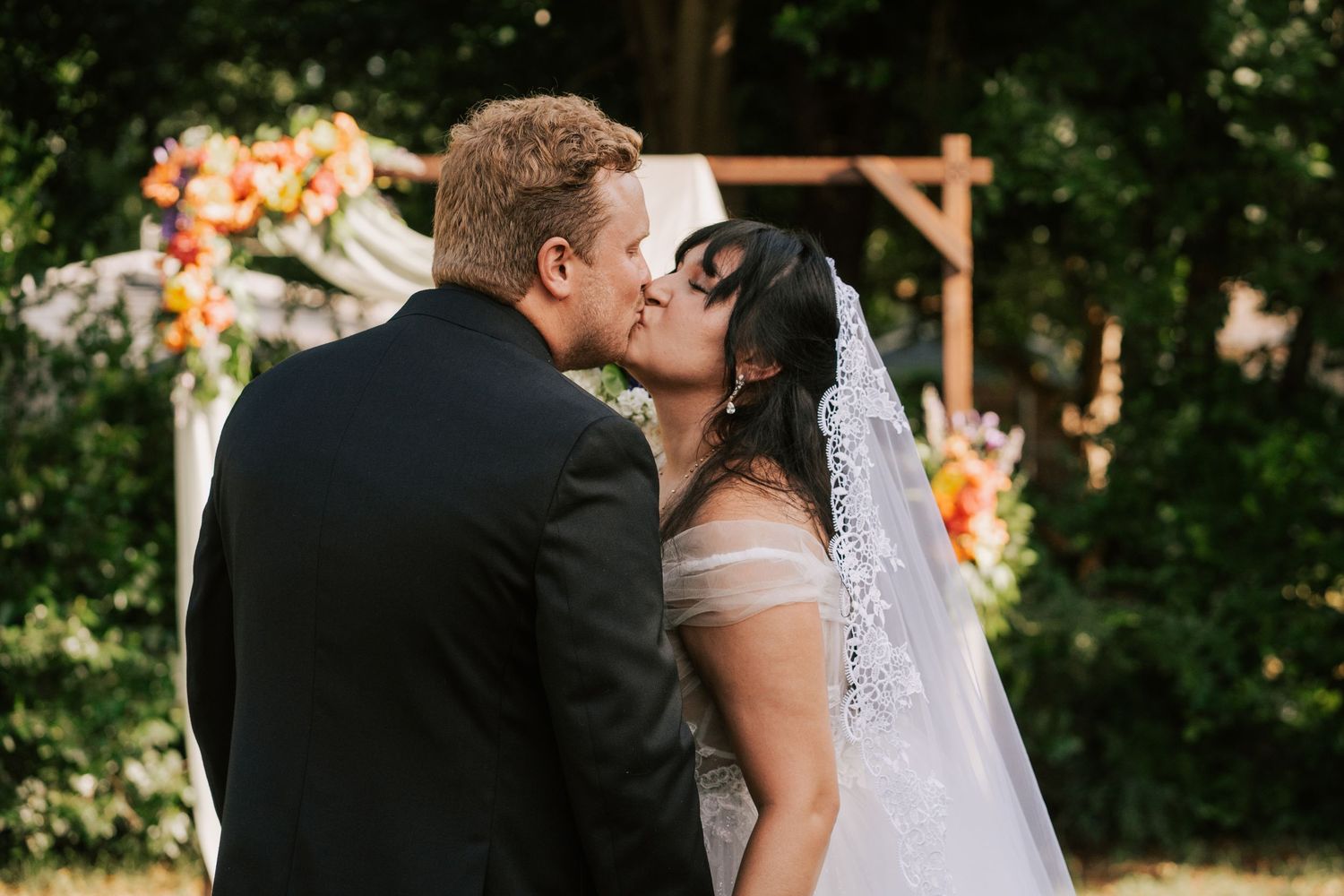 Bride and groom share their first kiss at an outdoor wedding ceremony with a floral arch backdrop.