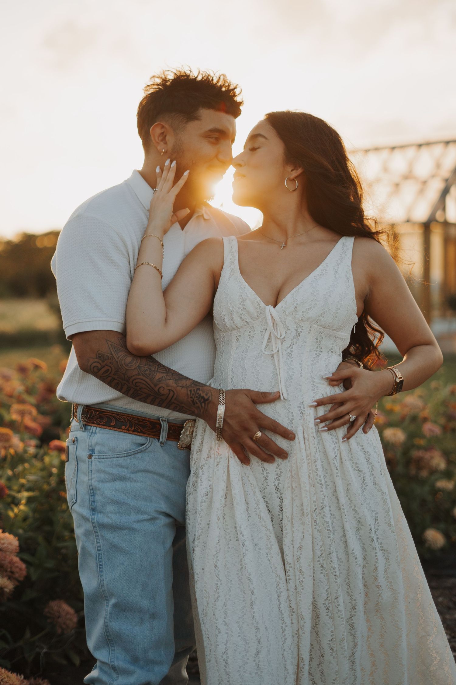 Couple sharing a romantic kiss at golden hour in a flower field, woman in white lace dress cradling baby bump.