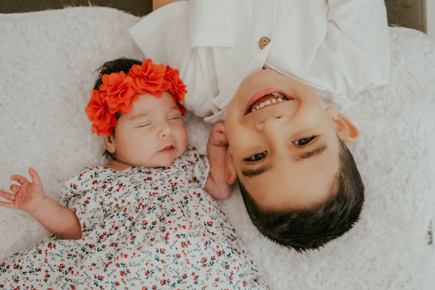 Newborn baby girl in floral dress with red flower headband lying next to smiling older sibling on white fur blanket.