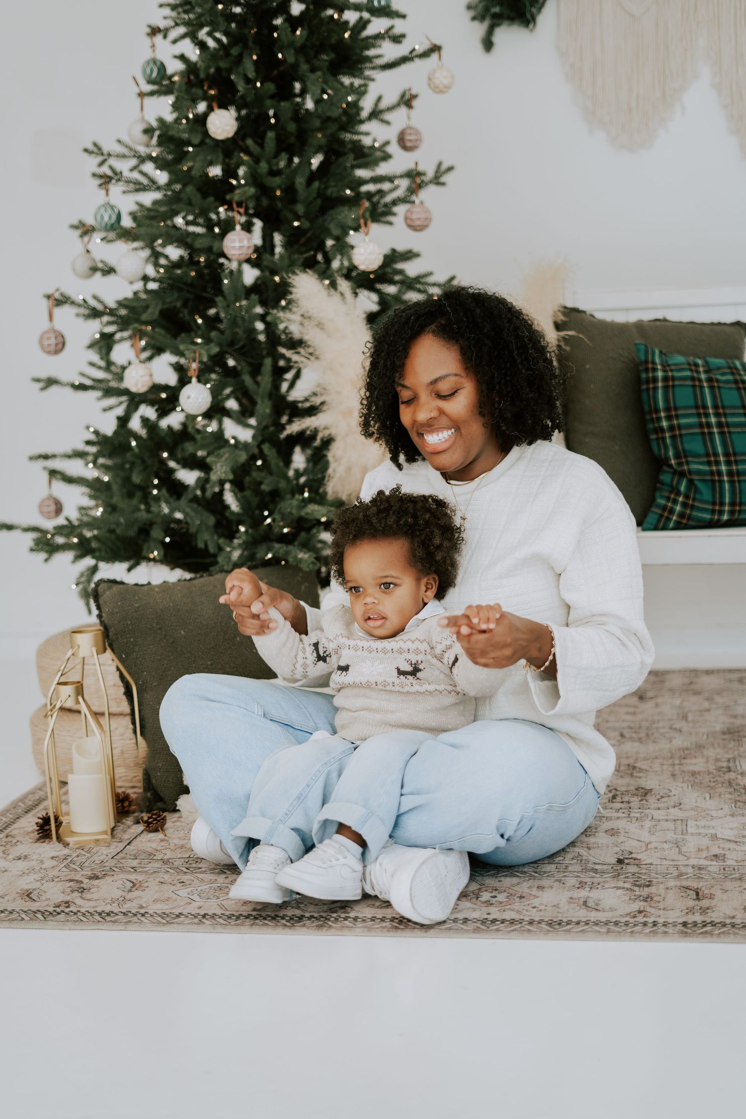 A happy mother sits on a rug with her toddler in front of a Christmas tree with pink ornaments.