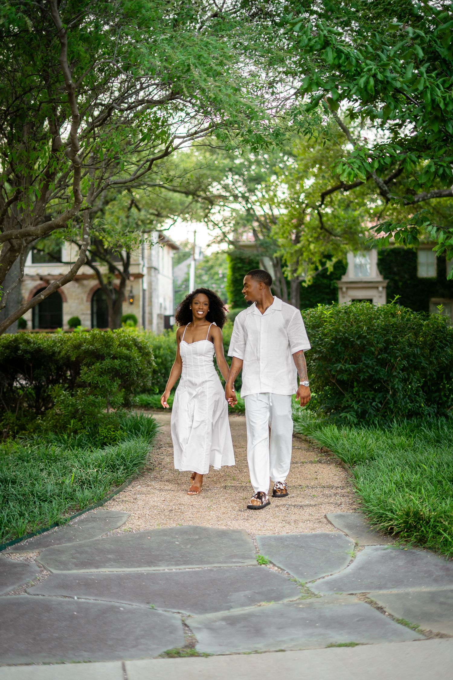 A stylish Black couple in white outfits walks hand-in-hand along a tree-lined garden pathway.
