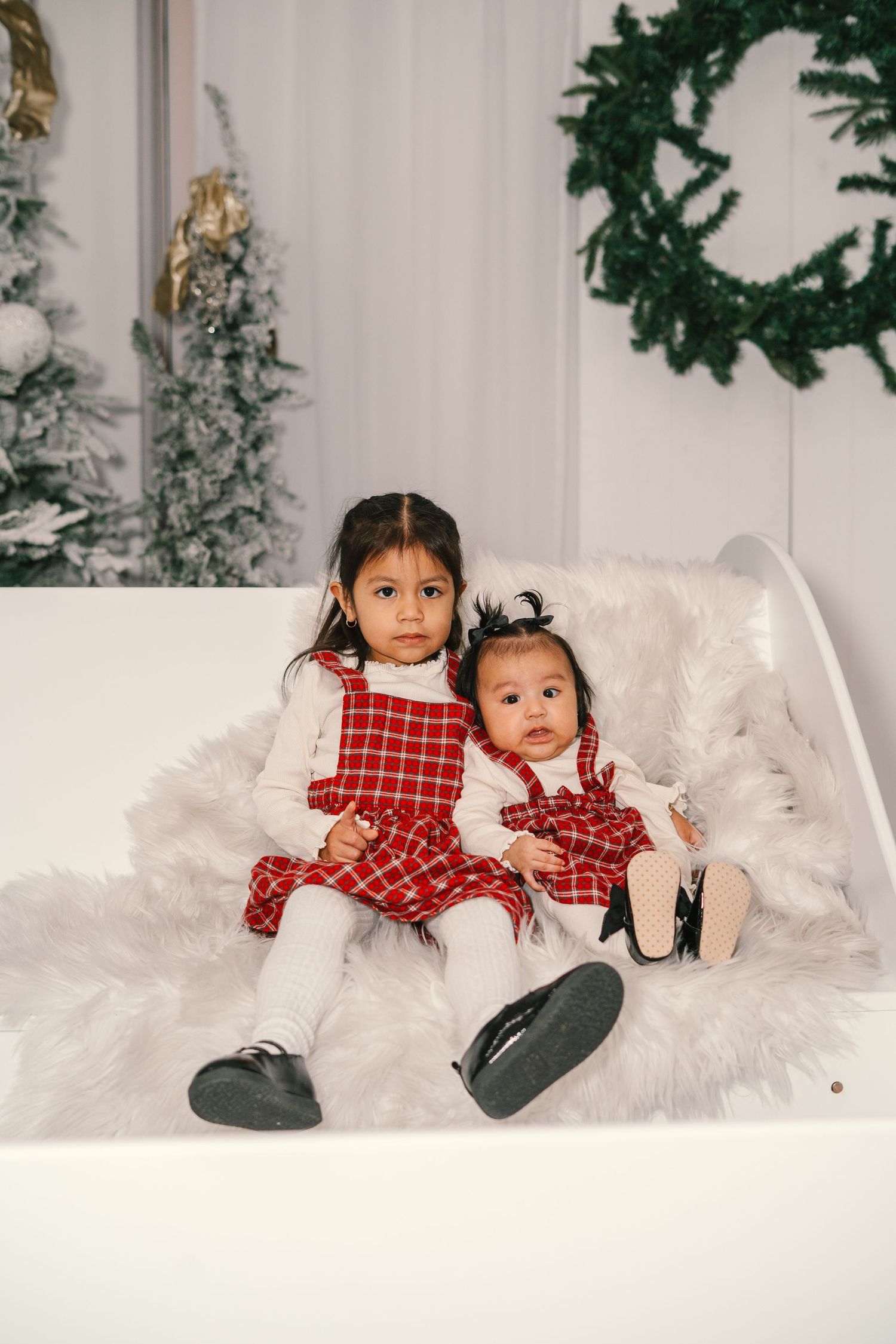Two young sisters in matching red plaid dresses pose together on a white fur blanket with Christmas decorations.