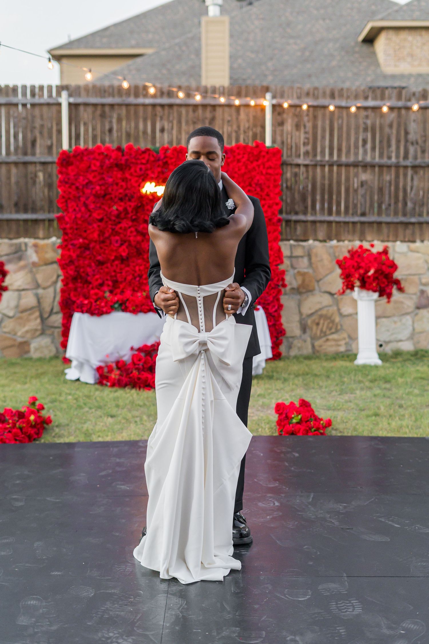 Bride and groom share first dance on outdoor stage with red rose backdrop at backyard wedding.