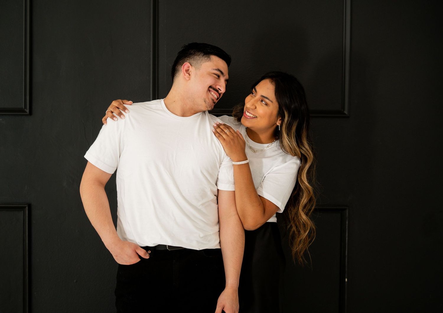 Happy couple in white t-shirts embracing and laughing against a dark paneled wall.