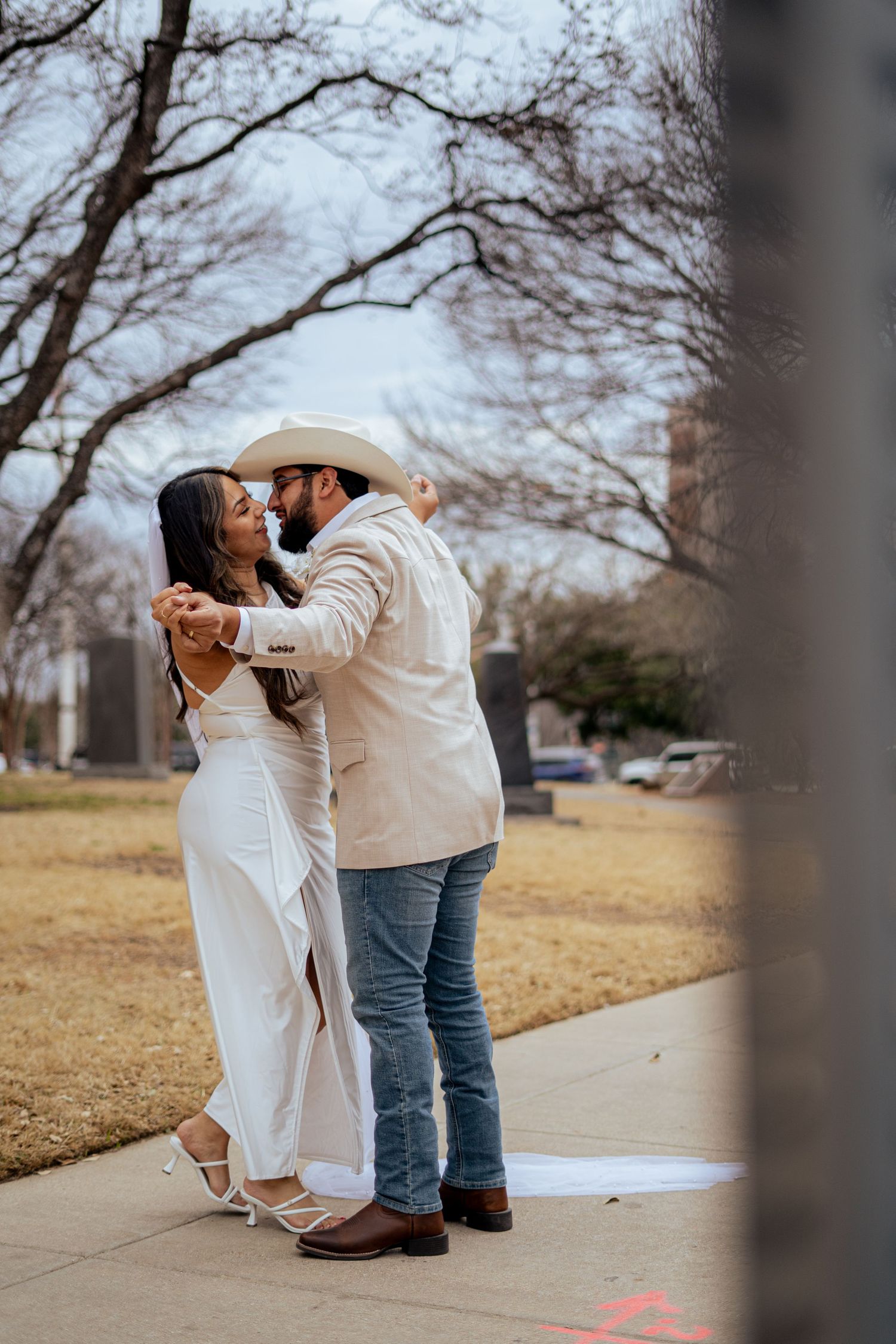 Newlywed couple dancing together on a sidewalk in western wedding attire with bare winter trees.