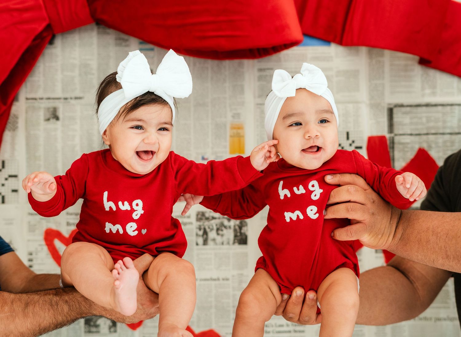 Two smiling babies in matching red 'hug me' onesies with white bows.