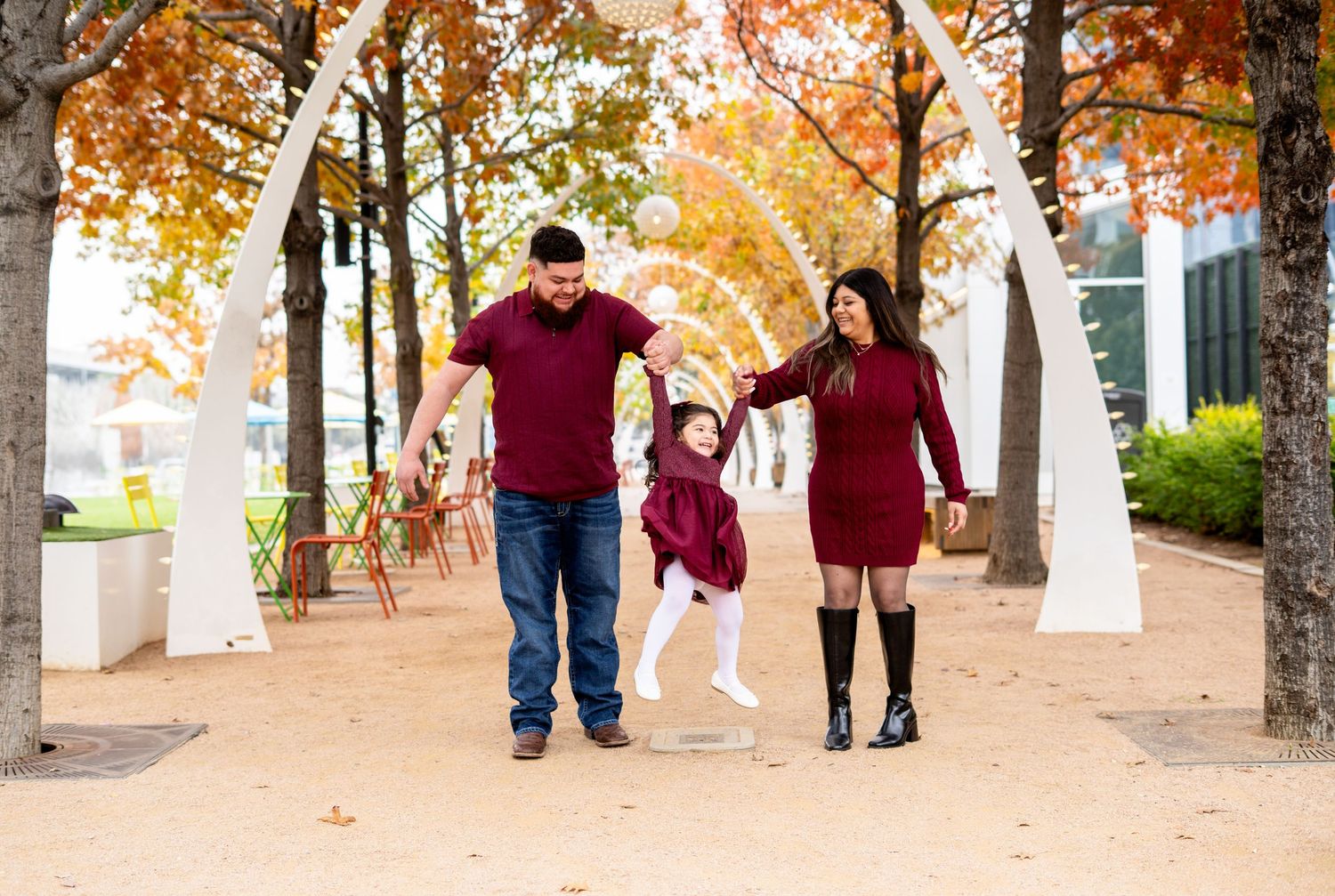 Parents swinging toddler daughter between them along a tree-lined autumn path with white arches.