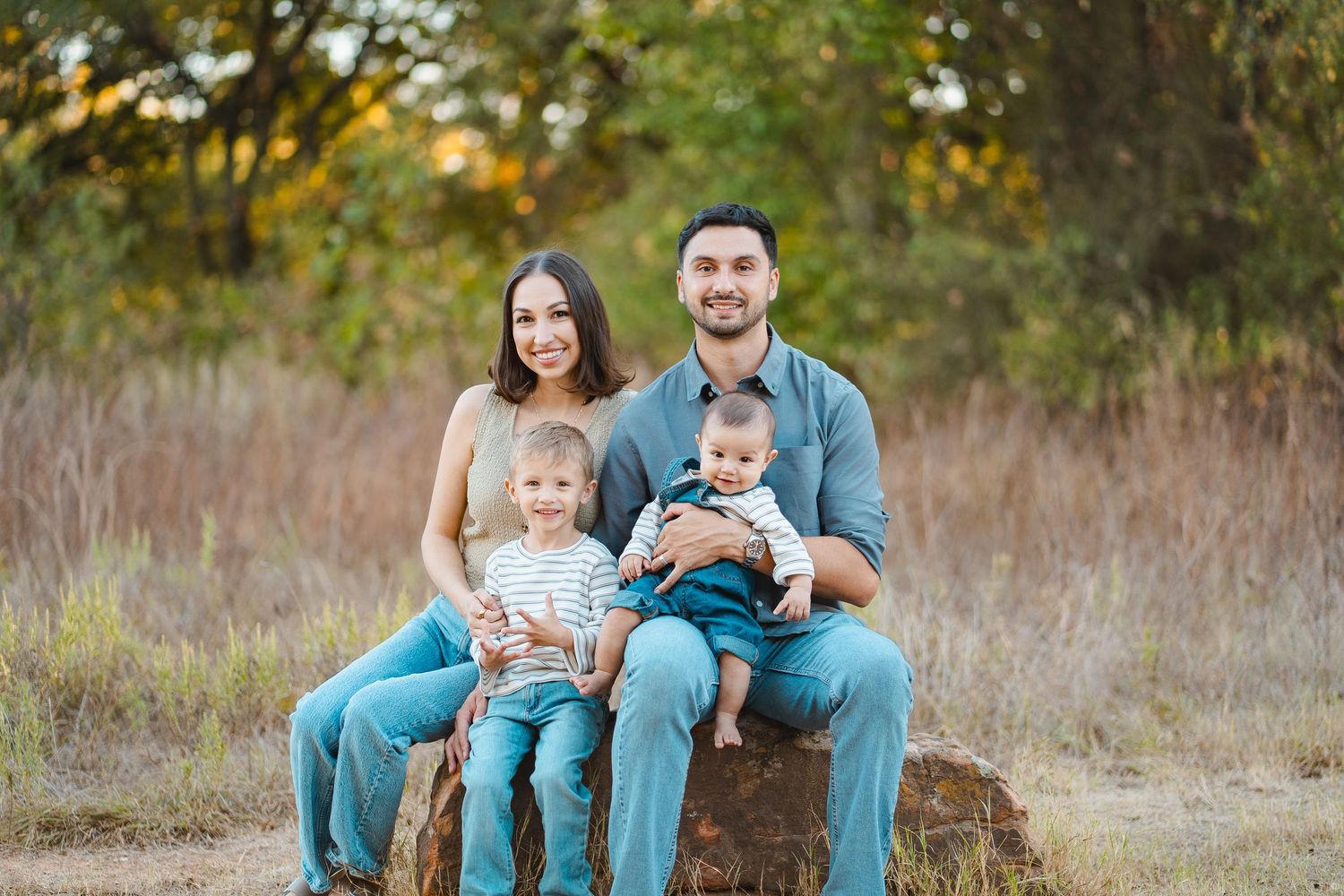 Happy family of four posing outdoors on a rock, parents with two young children in a sunny field.