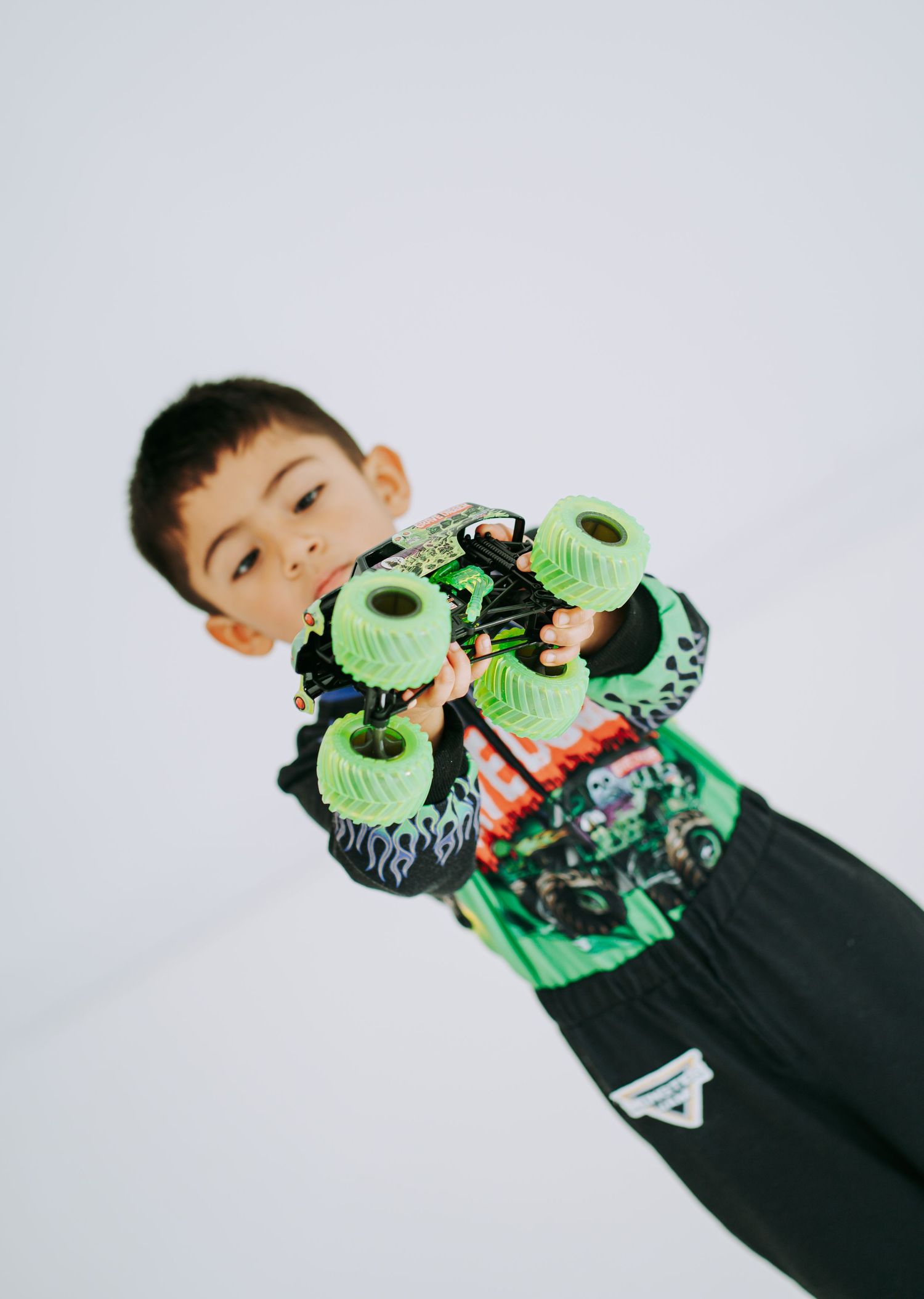 Child in Monster Jam outfit holds green Grave Digger monster truck toy toward camera on white background.
